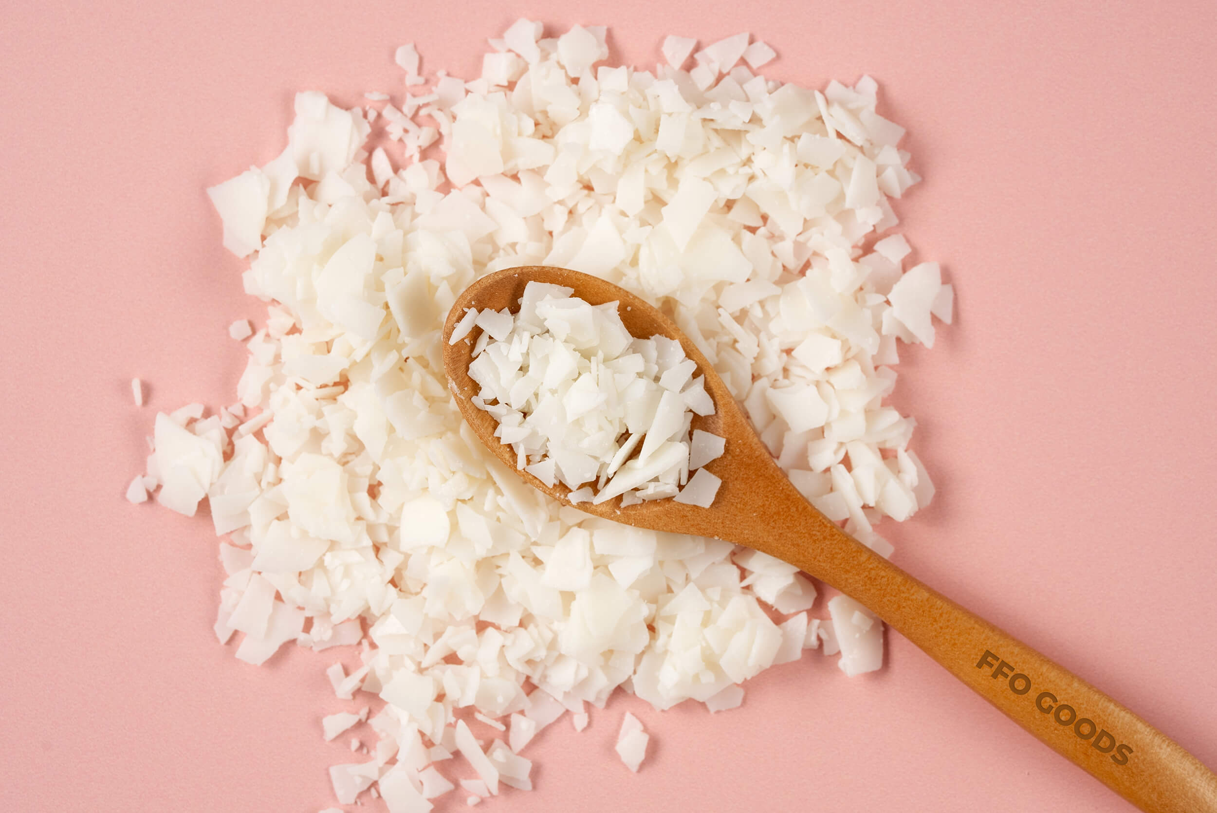 A wooden spoon filled with white flakes rests on a pile of similar flakes, placed against a pink background. The spoon has FFO GOODS inscribed on it.