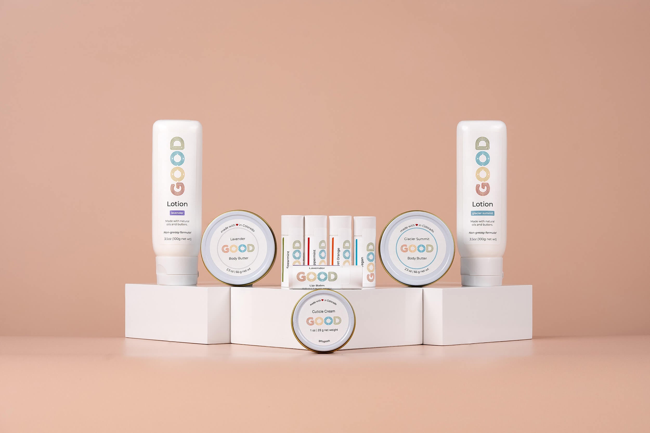 A tin of cuticle cream labeled GOOD is open next to a stick of lip balm with similar branding. They are placed on a wooden slab, surrounded by green leaves and purple flowers, against a beige background.