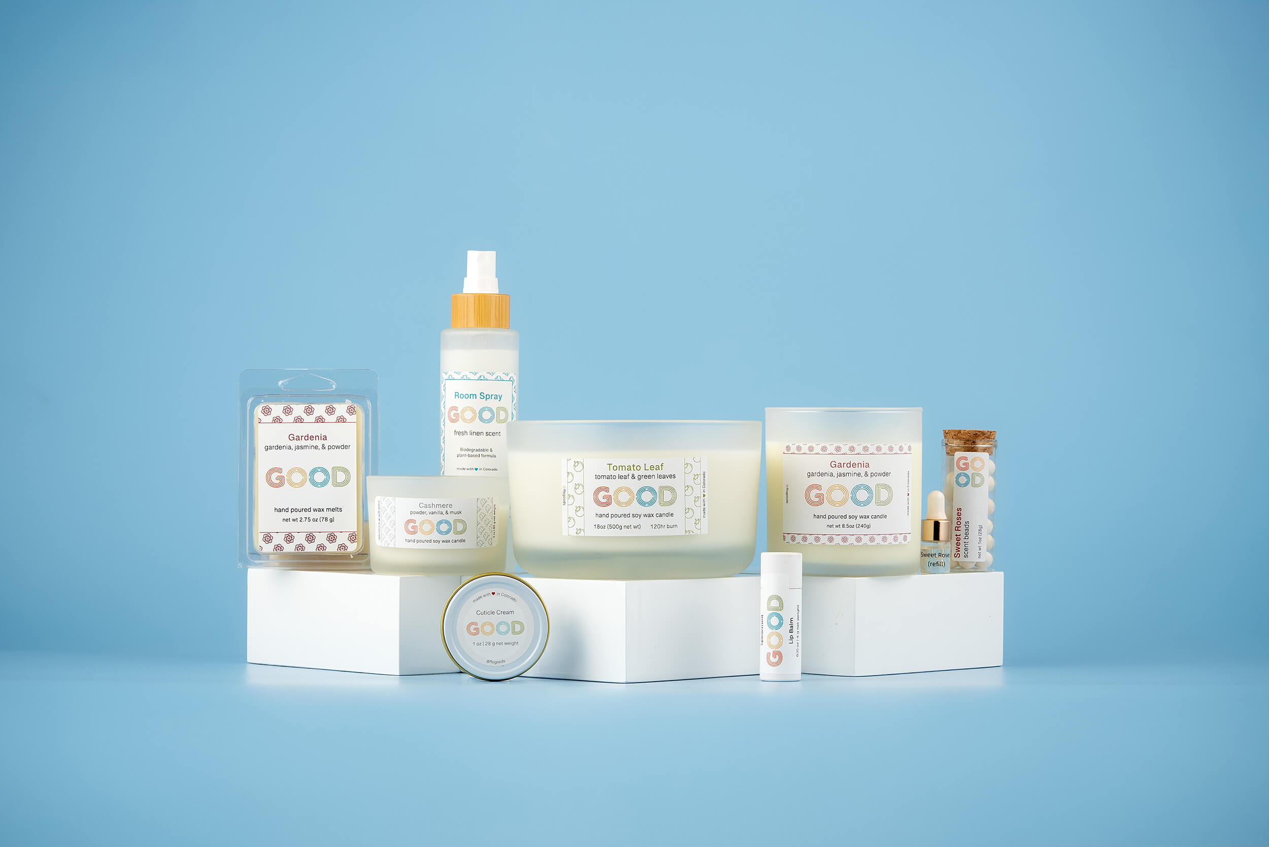 A display of candles and skincare products labeled "GOOD" arranged on pieces of wood against a pink background.