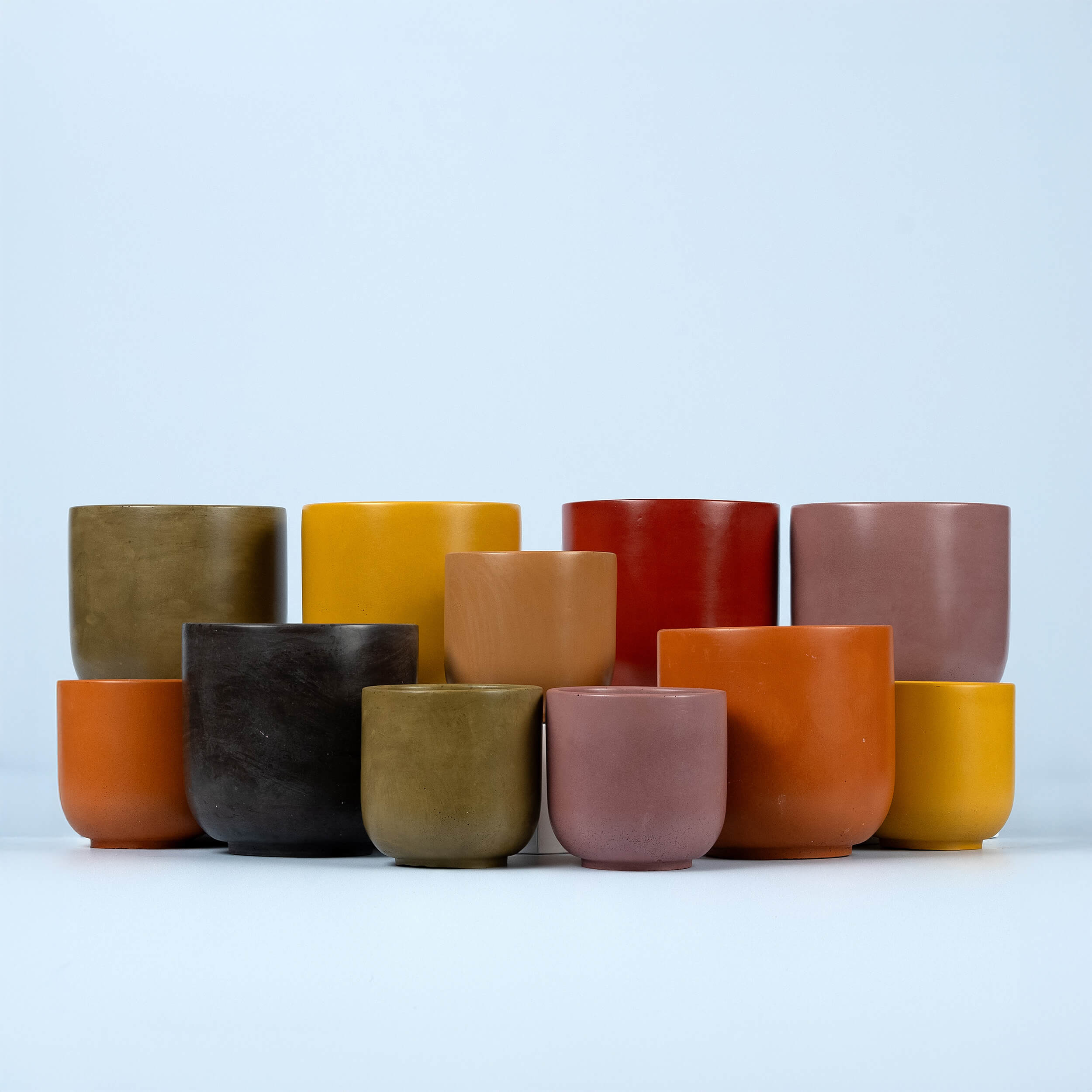 A group of twelve small, round plant pots in various earthy colors, including yellow, orange, red, brown, and olive green, arranged in two rows against a plain light blue background.