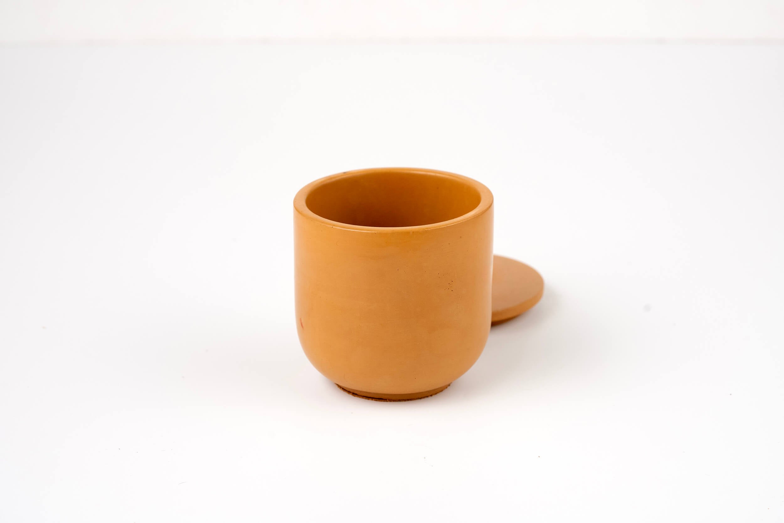 A small, round, brown Wheat Buff Candle Vessel with a matching flat lid behind it is displayed against a plain white background.