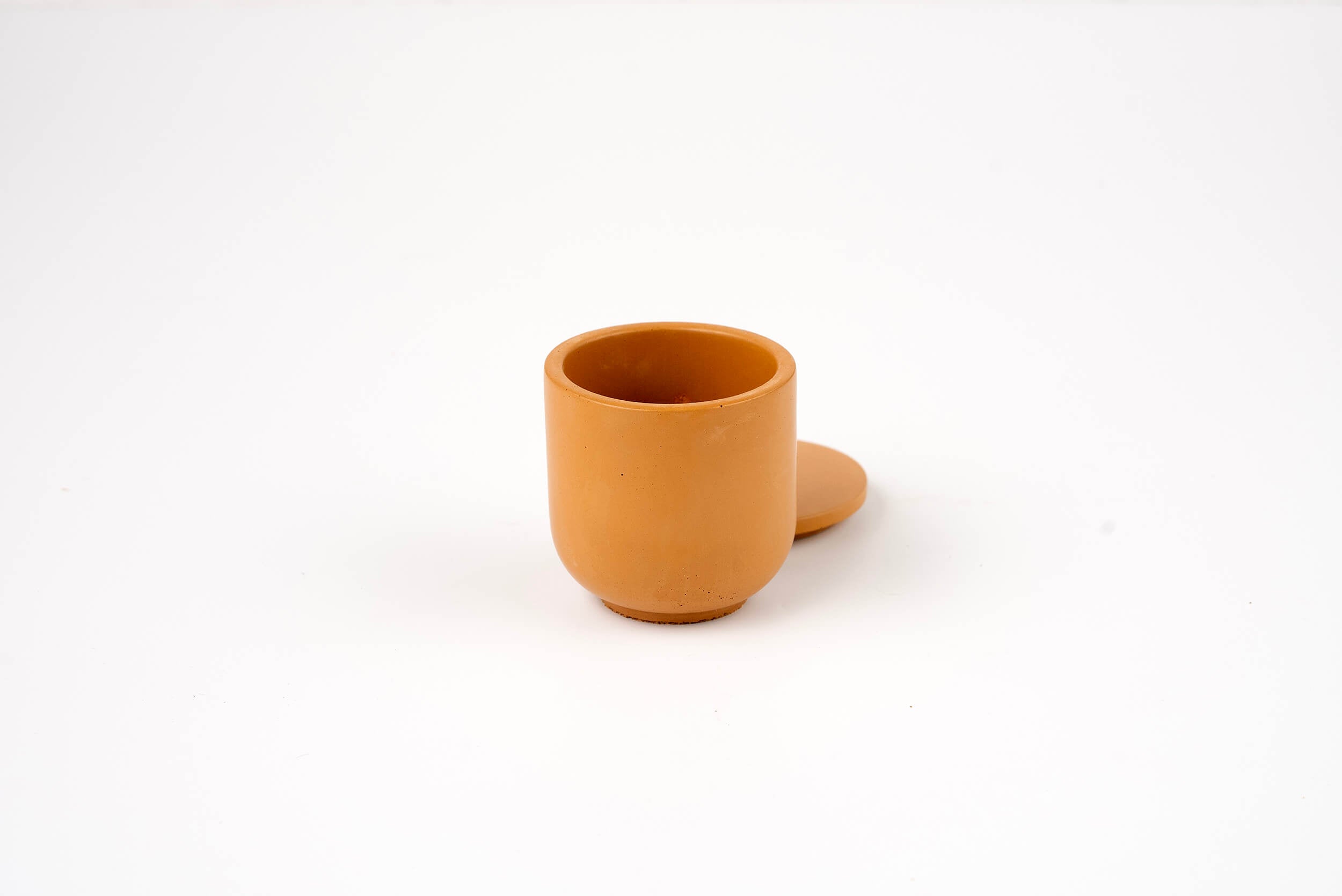 The Wheat Buff Candle Vessel, a small round brown clay cup with a matching saucer behind it, is displayed against a plain white background.