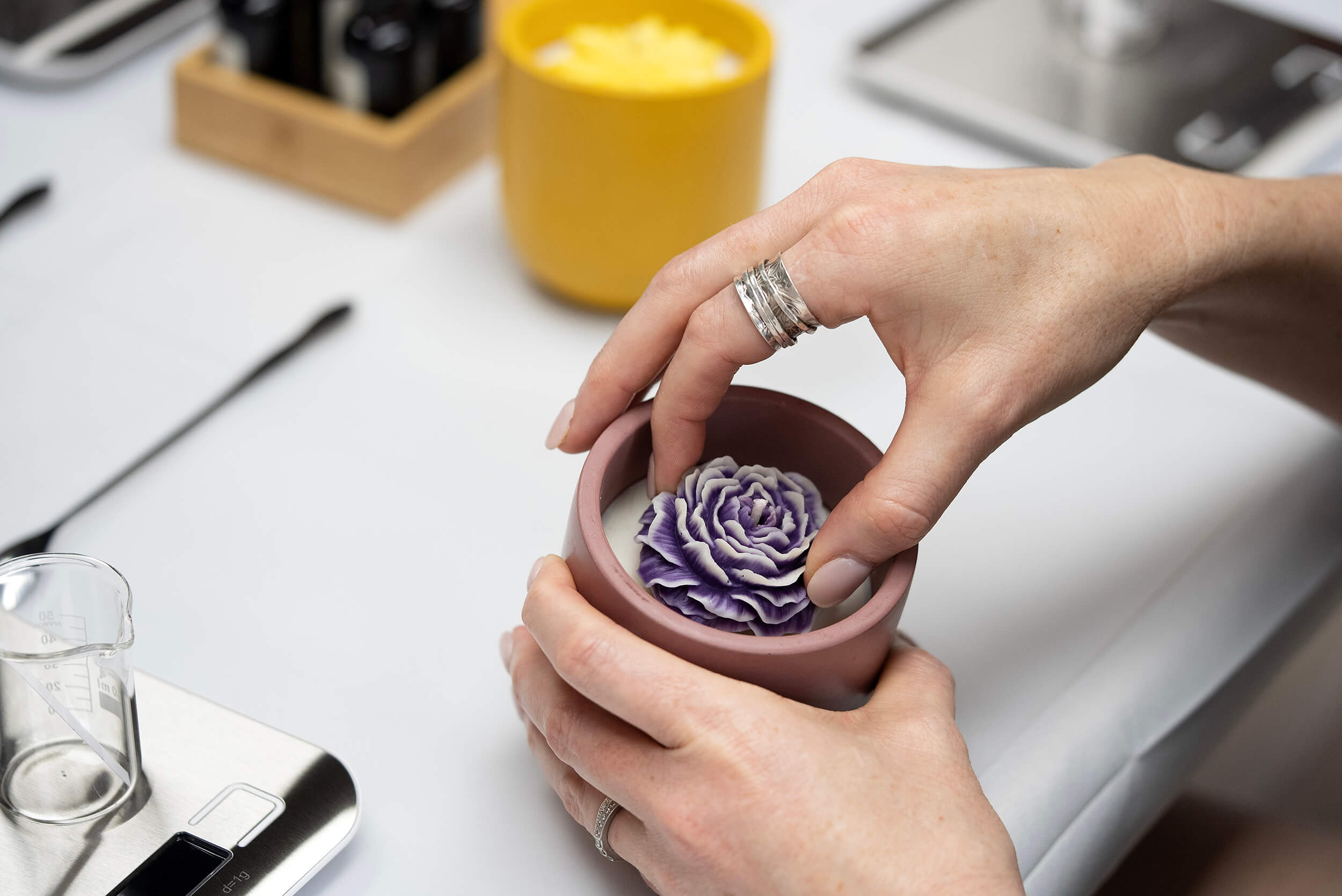 During the Candle Pouring Workshop, someone with rings places a purple and white artificial flower into a small pink pot. Scales, a beaker, and a yellow container are also visible on the white table in the background.