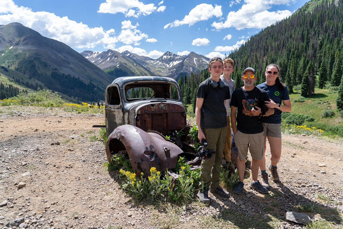 A family of four stands beside a rusty, abandoned car in a mountainous landscape. The scene includes rolling green hills, yellow wildflowers, and clear blue skies. Snow-capped peaks rise in the background.