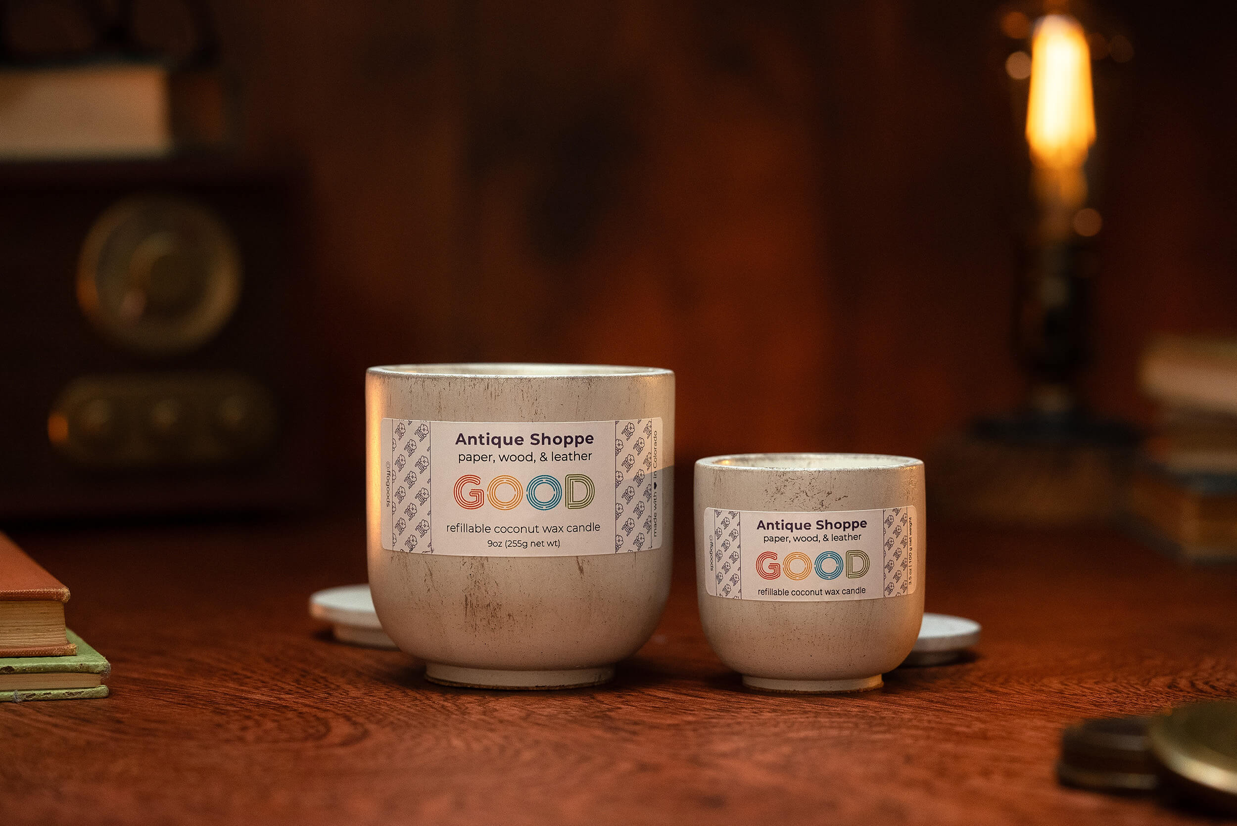 Two “Antique Shoppe” beige candles in ceramic jars, labeled with notes of paper, wood & leather, rest on a wooden table alongside vintage books, a radio, and a glowing Edison bulb lamp for a warm, nostalgic setting.