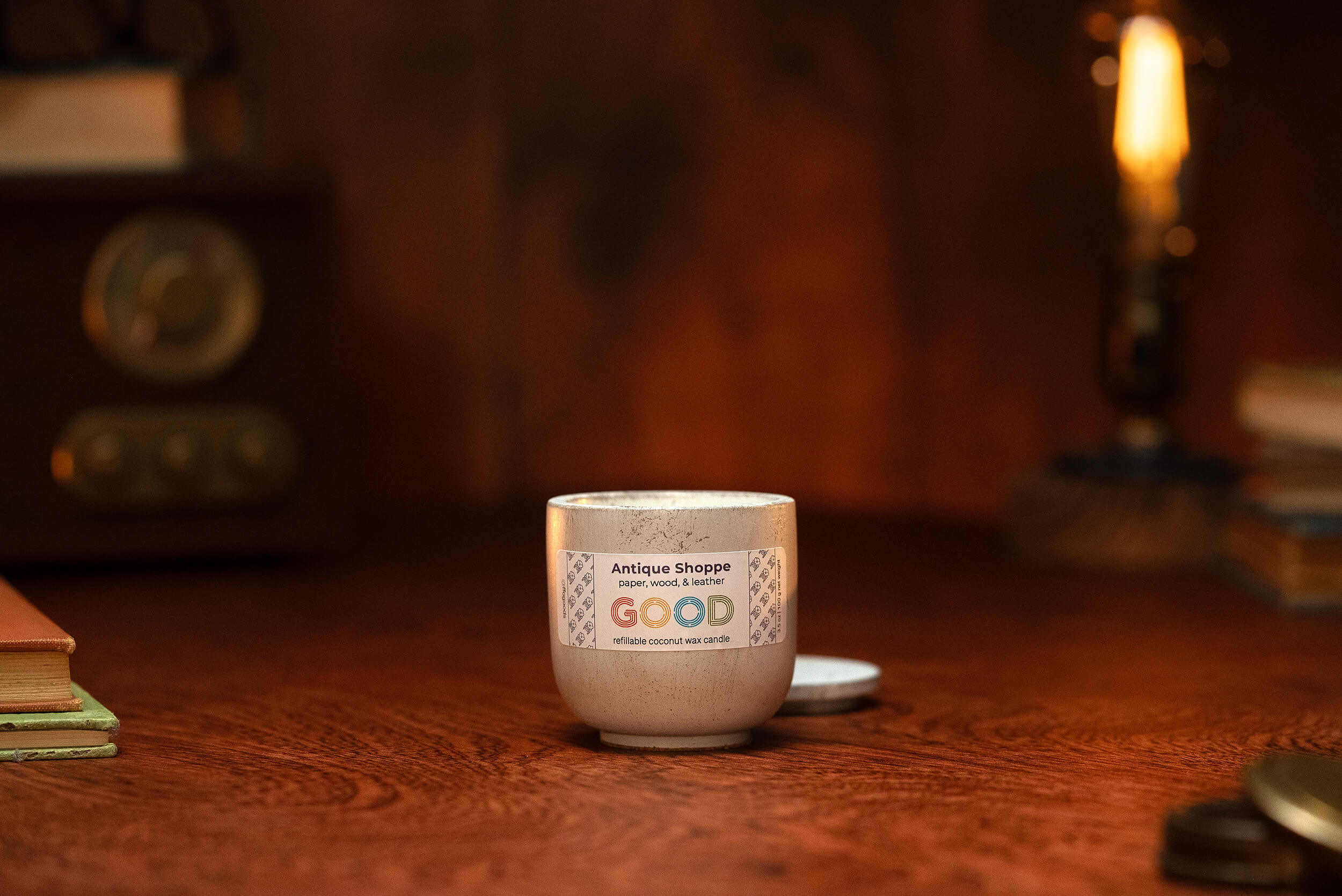 A lit Antique Shoppe candle sits on a wooden surface with books, a vintage radio, and a glowing bulb in the background, creating a cozy, warm atmosphere.