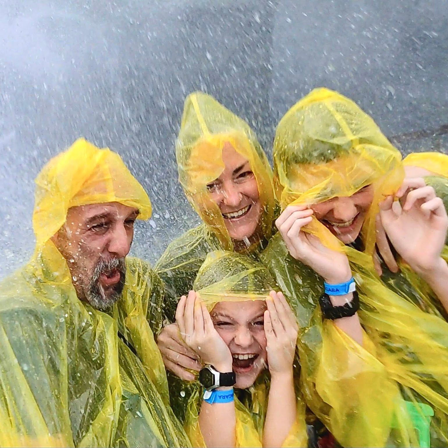 A group of four people, wearing yellow rain ponchos, laugh and shield themselves from water splashes. They appear excited and are likely on a water ride or in a wet area, enjoying the moment together.