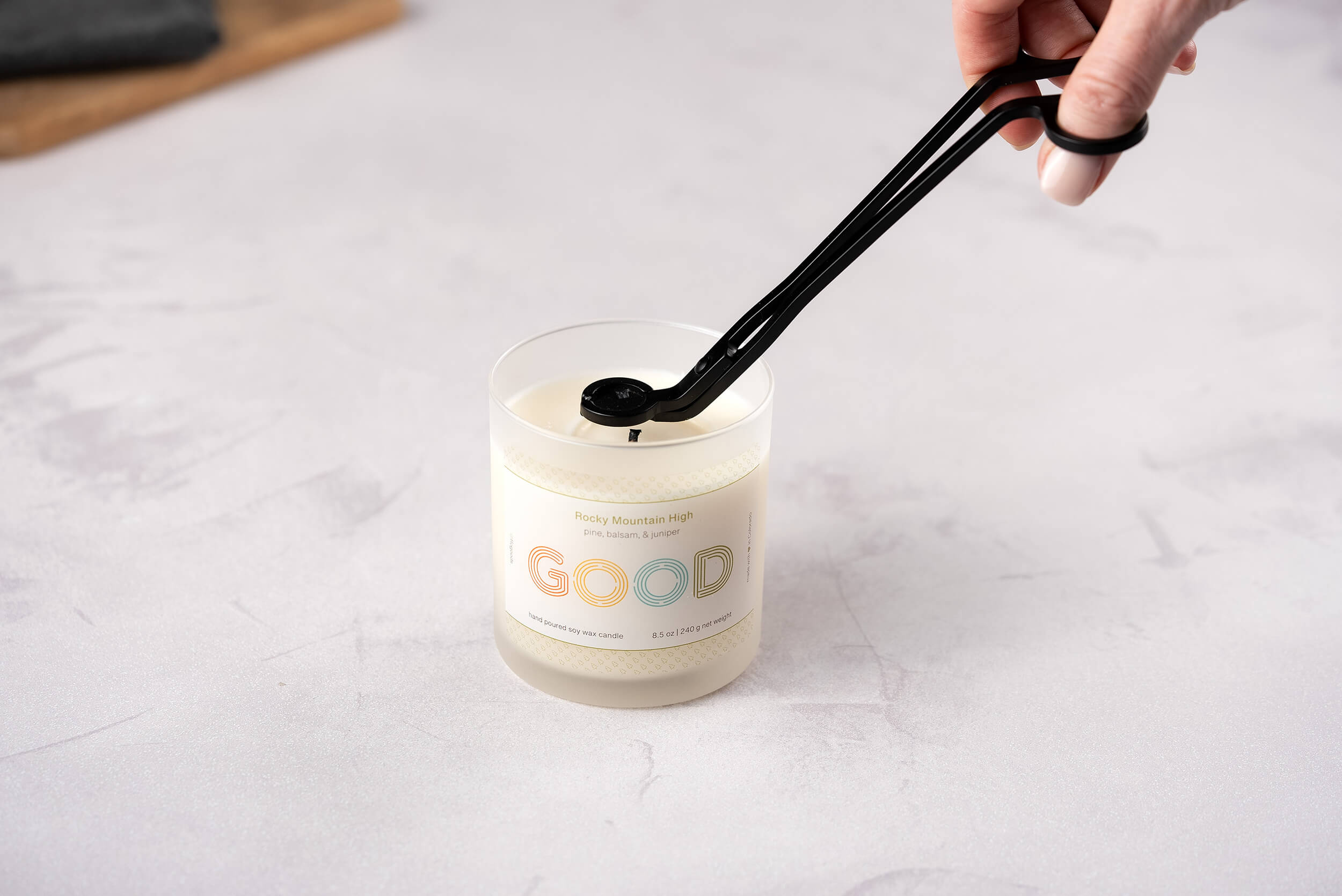 With expert precision, a hand uses the Wick Trimmer from FFO Goods to neatly shorten the wick of a candle marked with the label GOOD. This meticulous act of maintenance ensures that the white candle, elegantly placed on a white surface, burns at its best.