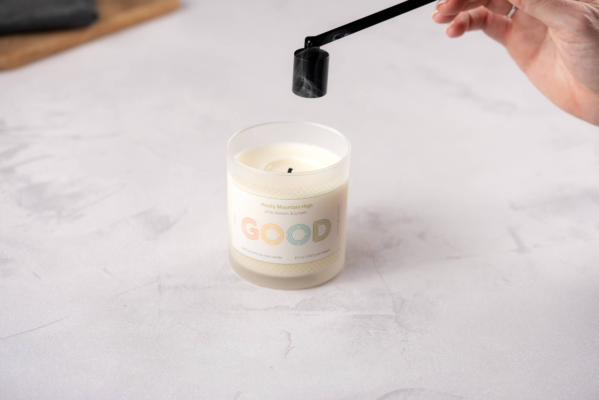 A hand elegantly holds a Candle Snuffer over a lit candle in a jar labeled FFO Goods. The durable construction of the snuffer ensures safety as it hovers above the flame, resting on a light gray surface.