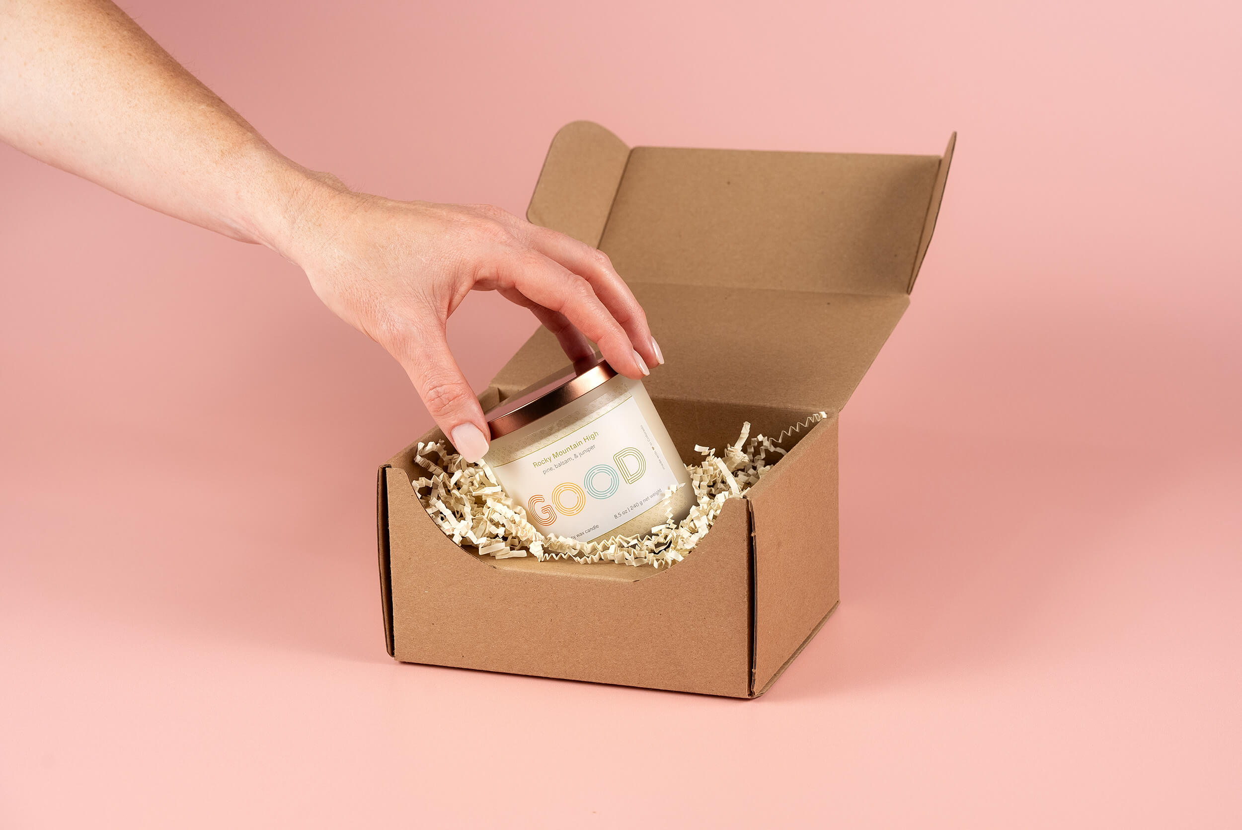A hand placing a white candle with a rose gold lid labeled GOOD into a brown cardboard box filled with shredded paper, set against a pink background.