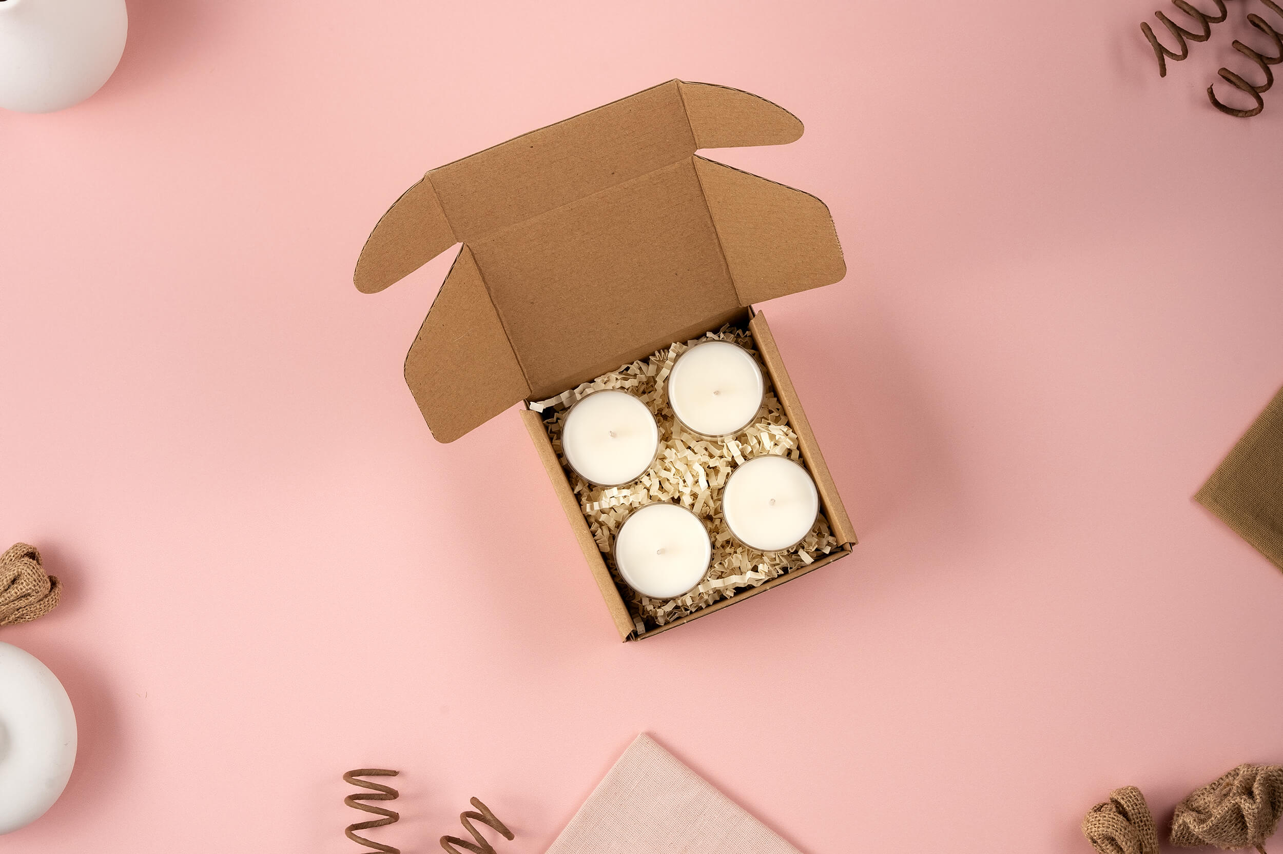 A soft pink background sets the stage for a cozy, minimalist scene featuring an open cardboard box from FFO Goods containing four "Sample Sniffs" white tea-light candles nestled in shredded paper, surrounded by decorative touches like twine and a fabric piece.