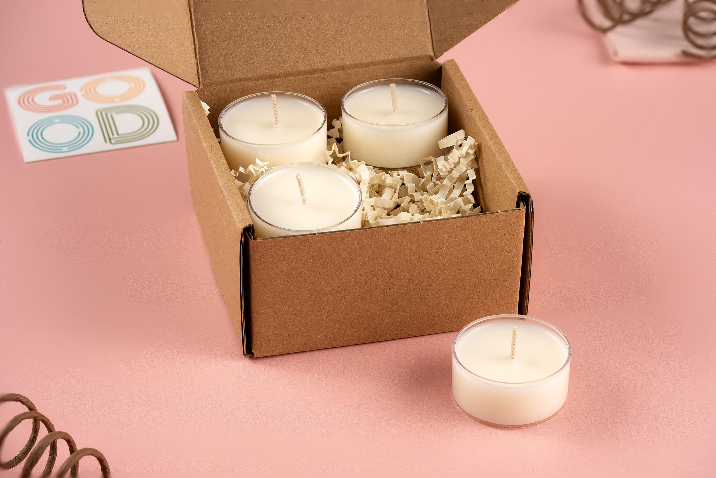 A cardboard box from FFO Goods contains four Sample Sniffs tea-light candles nestled within shredded paper. One candle is placed outside the box on a pink surface, while a card displaying the word GOOD in colorful letters suggests a delightful scent variety.