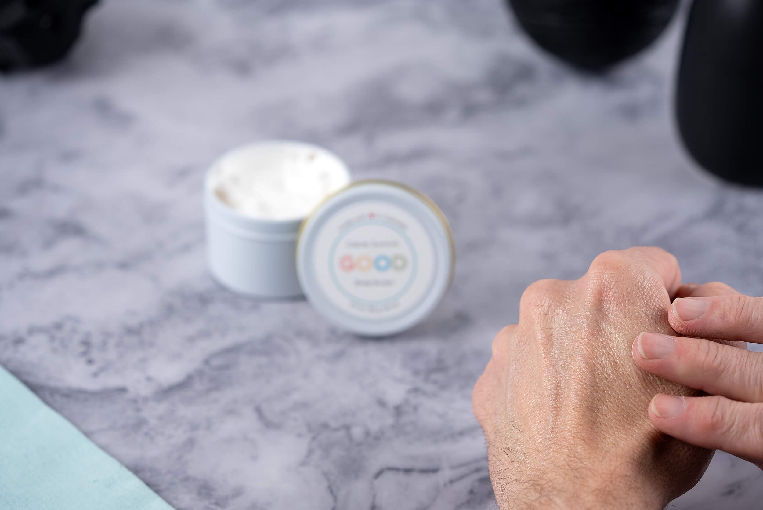 A person applies Whipped Body Butter to the back of their hand. In the background, an open white container of Whipped Body Butter sits on a marble-textured surface.