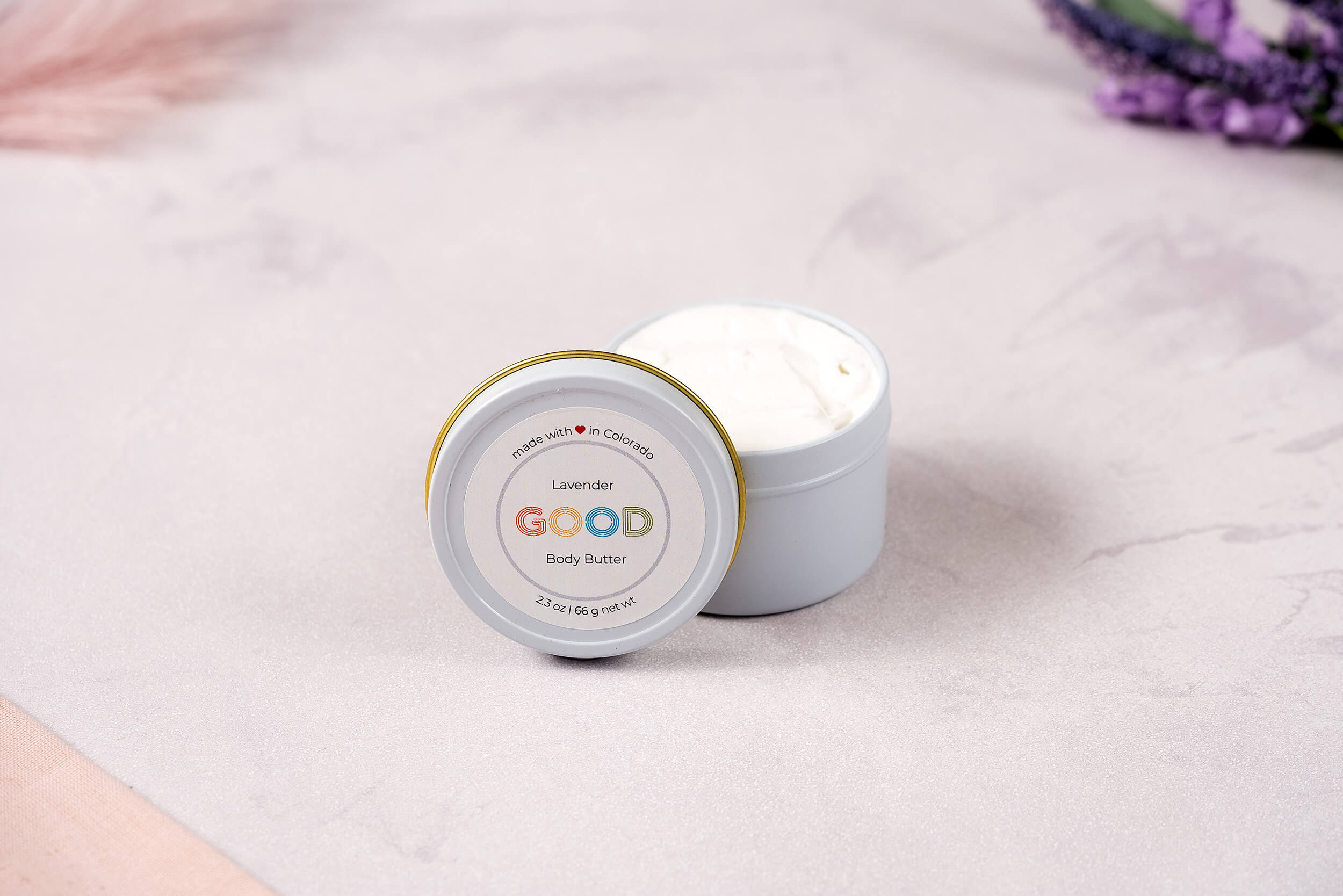 A small white container of Whipped Body Butter with its lid off reveals creamy, ultra-moisturizing lavender body butter. The label on the lid reads Lavender GOOD Body Butter. The background is light pink with purple flowers.