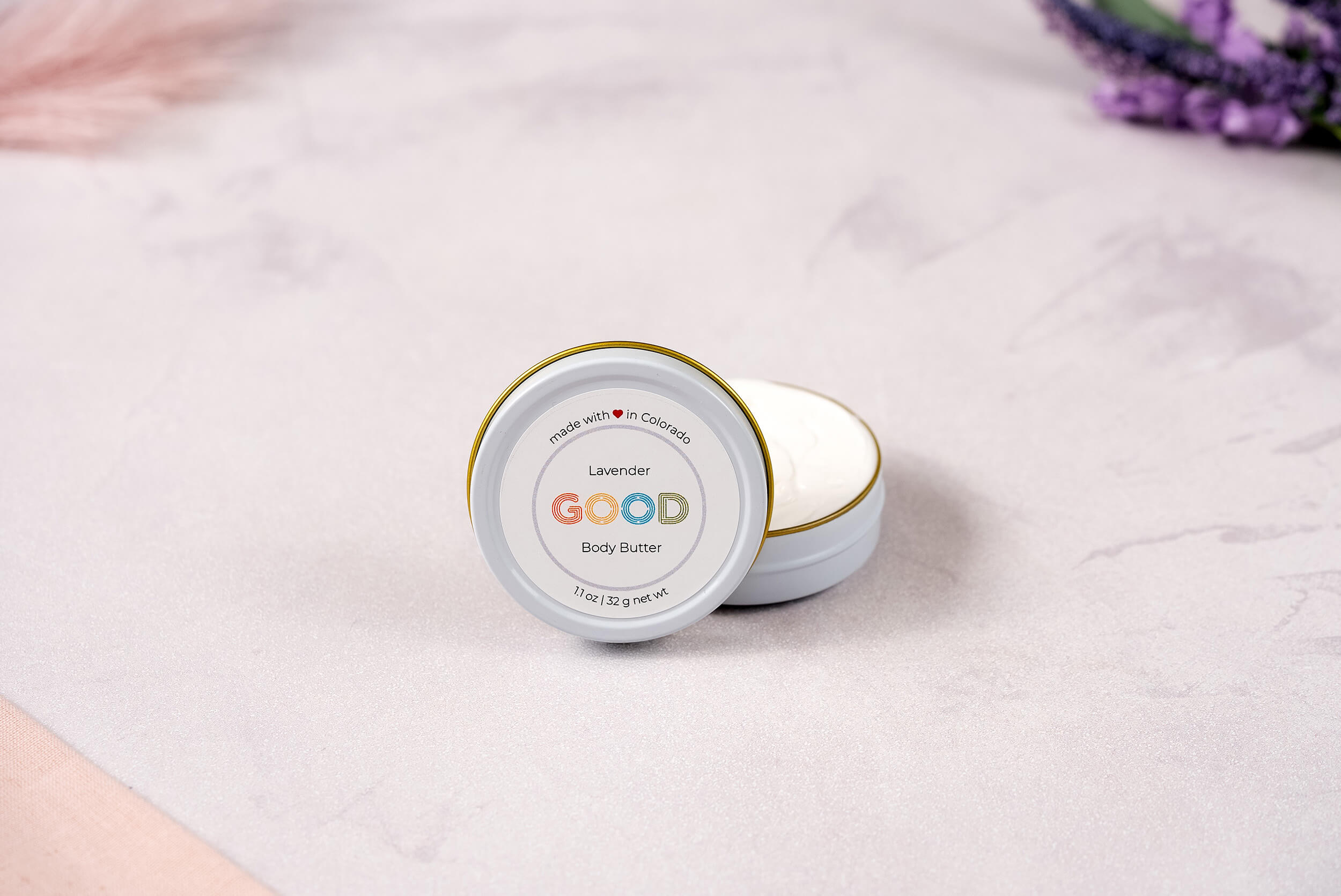 An open tin of Whipped Body Butter labeled GOOD rests on a light, textured surface with soft pink and purple accents in the background.