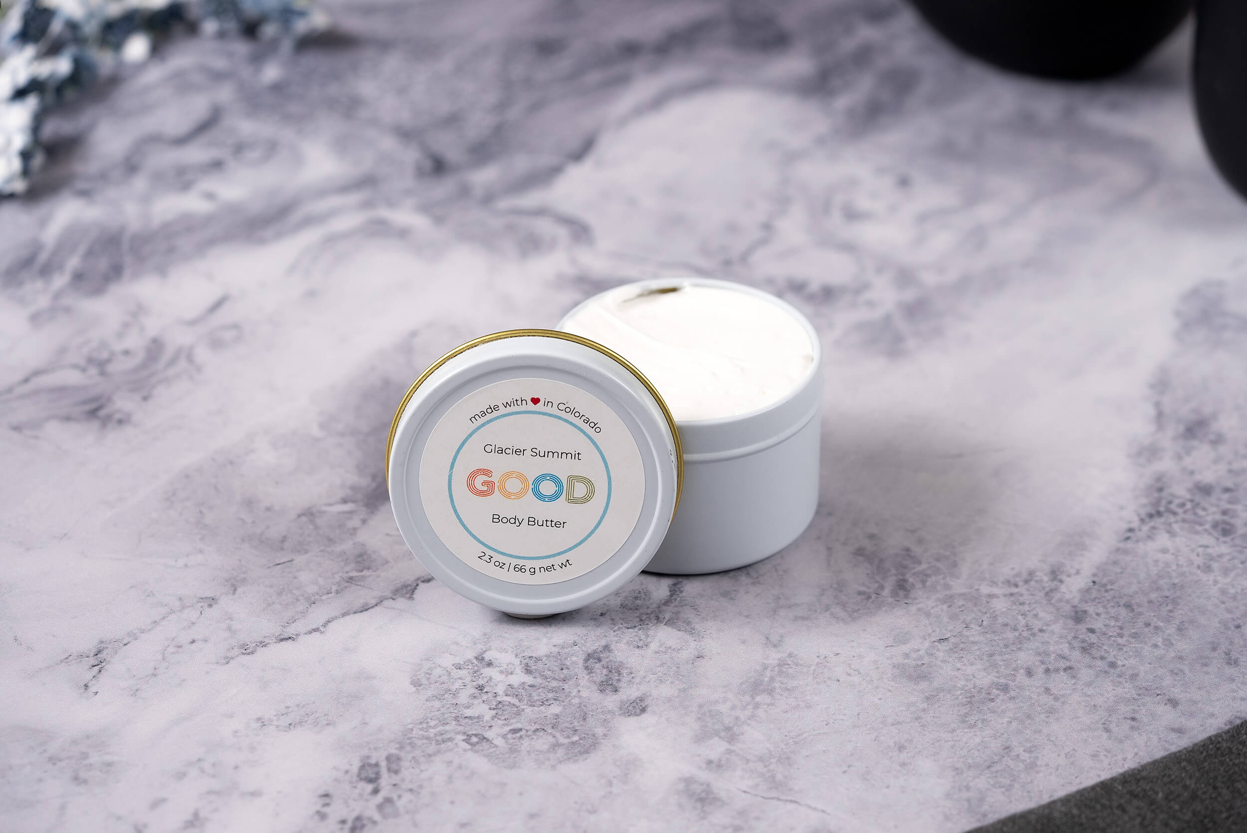 A small white jar of Whipped Body Butter sits open on a marbled surface, revealing rich cream inside. The visible lid label highlights its vegan skincare benefits and ultra-moisturizing formula.