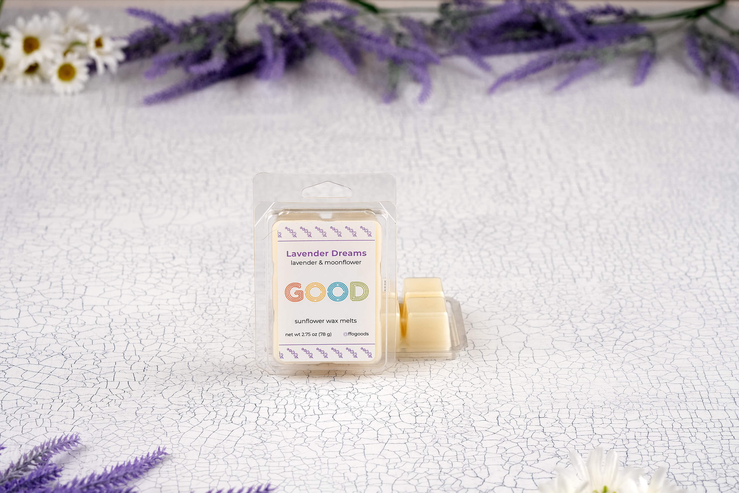 An open package of Lavender Dreams Wax Melts rests on a cracked white surface, with purple lavender and white flowers in the background. The label features a colorful, inviting design.
