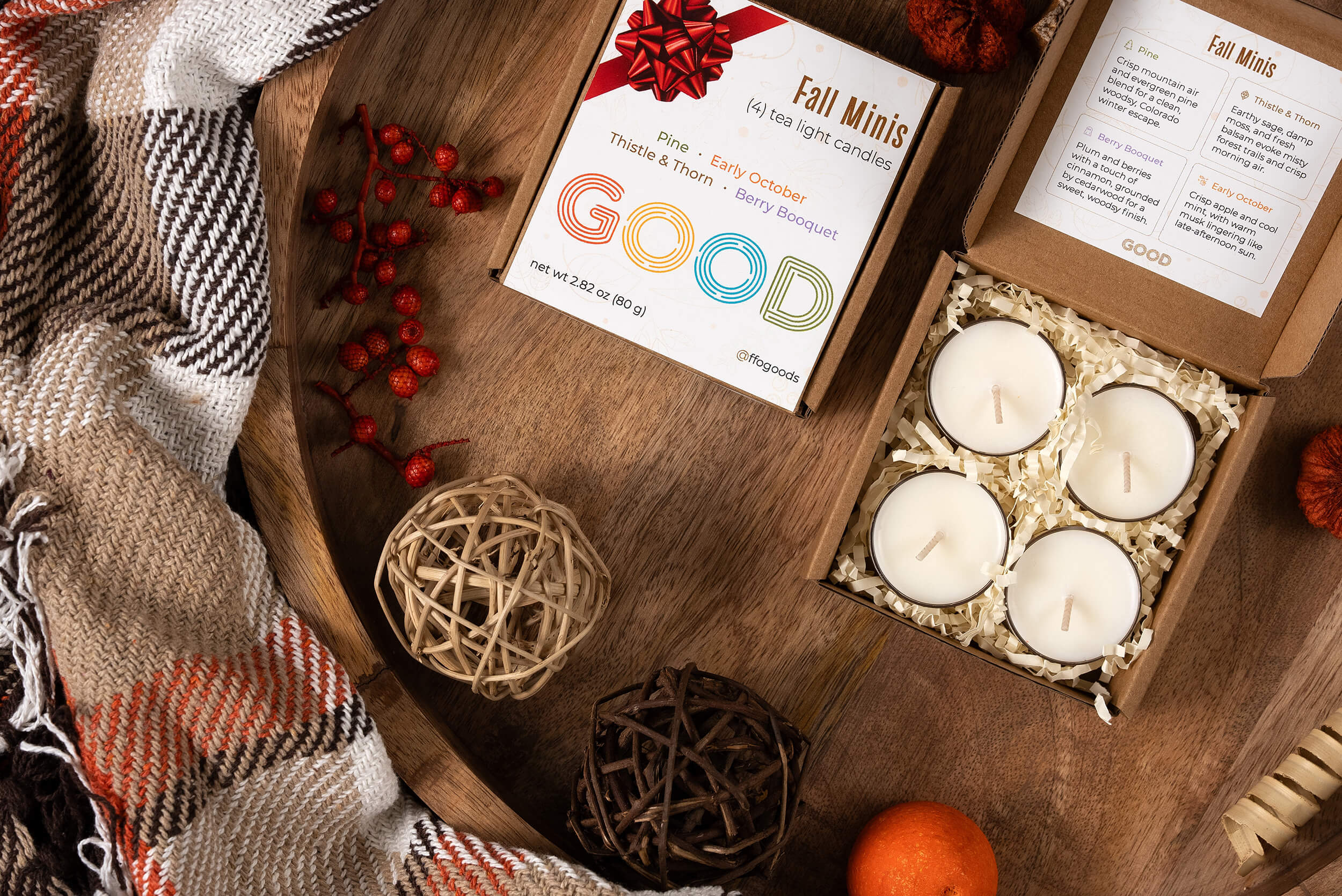 A cozy autumn flat lay with a plaid blanket, decorative balls, dried berries, an orange, and a box of four coconut wax Candle Minis on a wooden tray—perfect seasonal candles for fall.