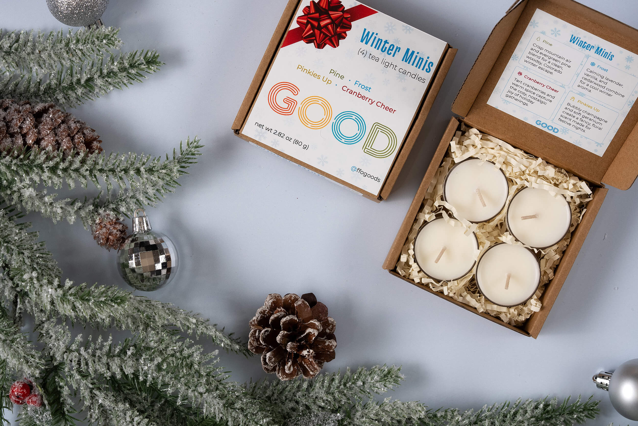 A box of four Candle Minis coconut wax tea lights is open on a light blue surface beside pine branches, a pinecone, and silver ornaments, creating a festive winter look with these seasonal candles.