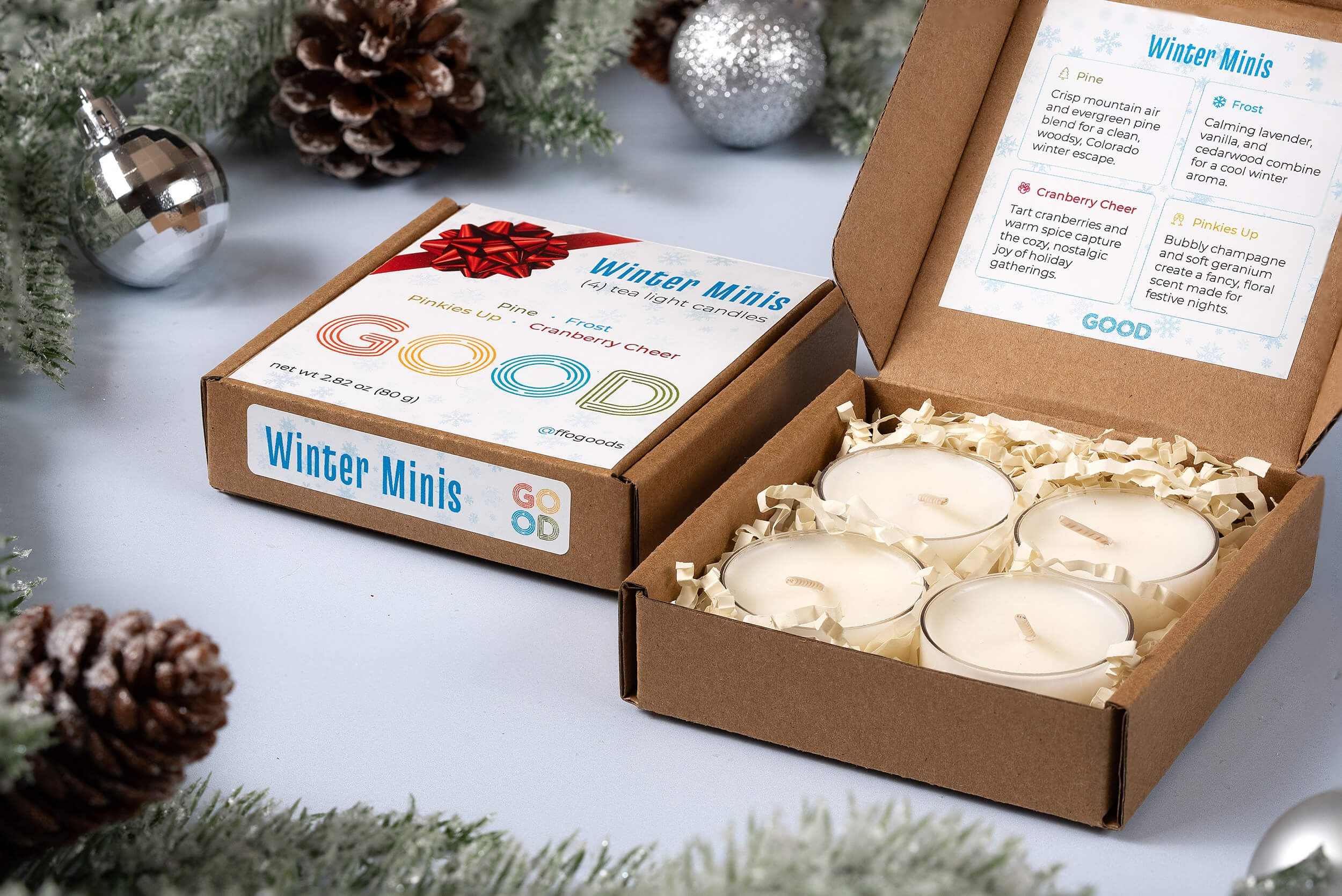 Four coconut wax Candle Minis in a brown box with shredded paper, next to the lid labeled Candle Minis and listing seasonal scents. Pine branches, pinecones, and ornaments create a festive winter scene in the background.