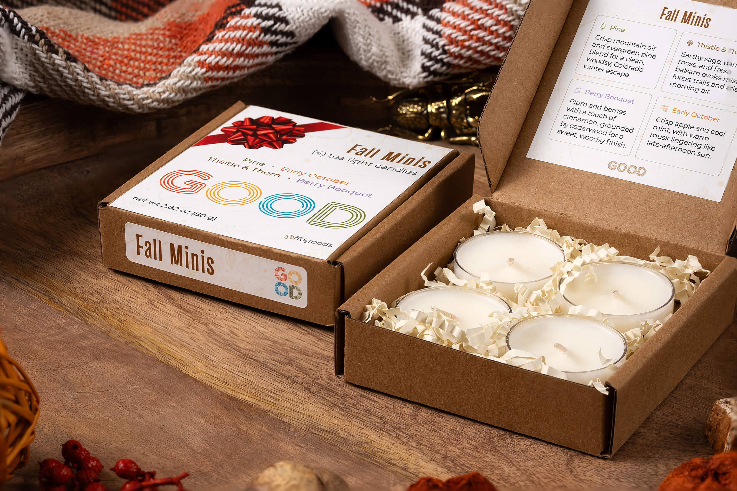 The Candle Minis box holds four white coconut wax tea lights in shredded paper. The open lid displays scent descriptions. Set on wood with fall décor and a plaid blanket, the brown box is labeled "Candle Minis.