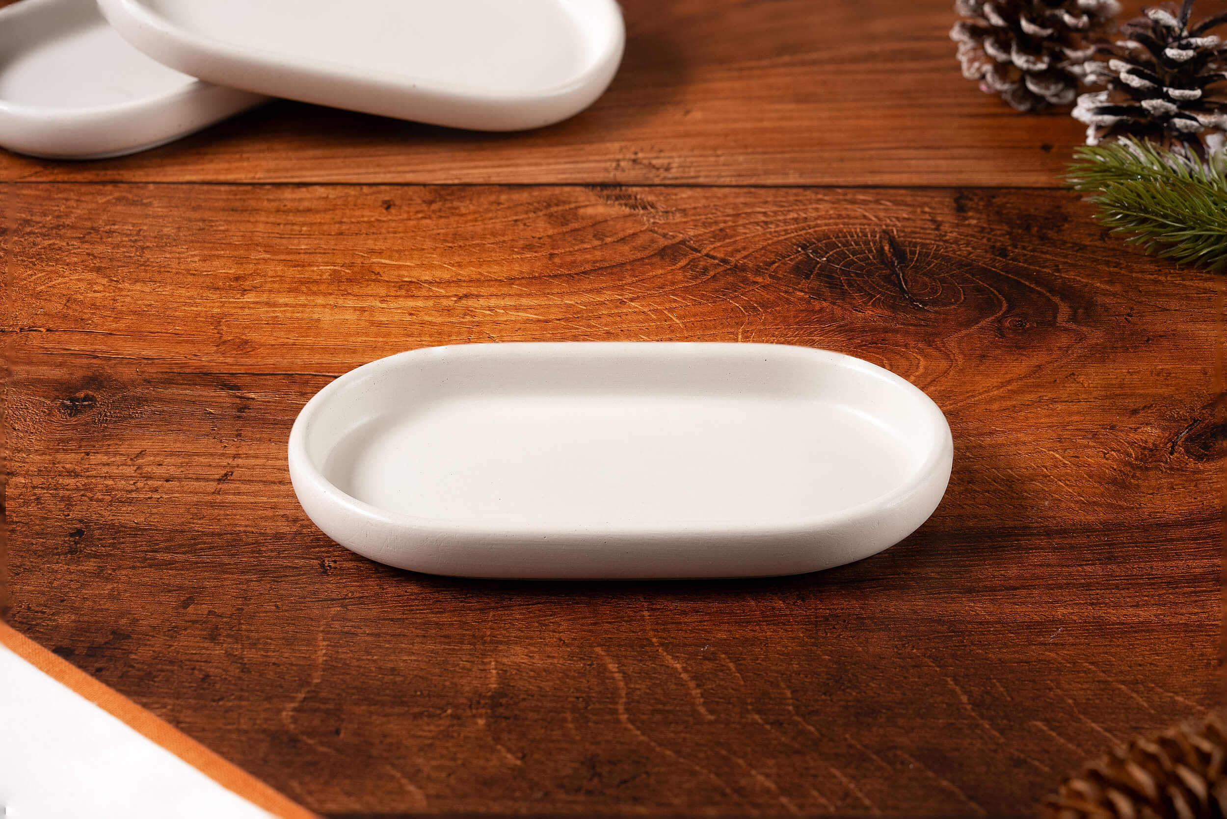 A small, white Concrete Candle Tray rests on a wooden surface with two stacked trays behind it and pinecones and evergreen branches in the background—ideal as a handcrafted, minimalist decor accessory for cozy spaces.