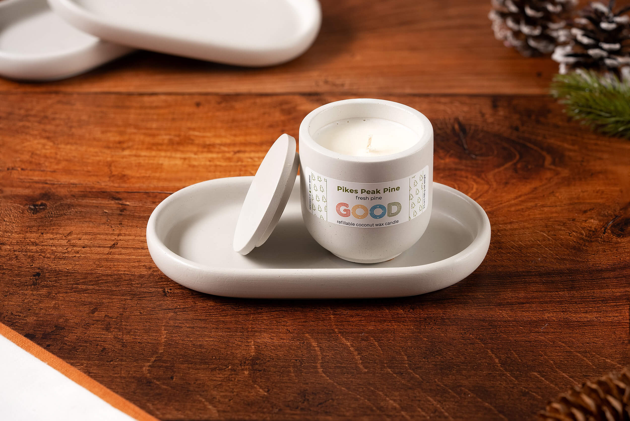 A white candle labeled Pikes Peak Pine with a partially open lid sits on a Concrete Candle Tray atop a wooden surface. Pinecones and greenery blur in the background, enhancing the minimalist home decor aesthetic.
