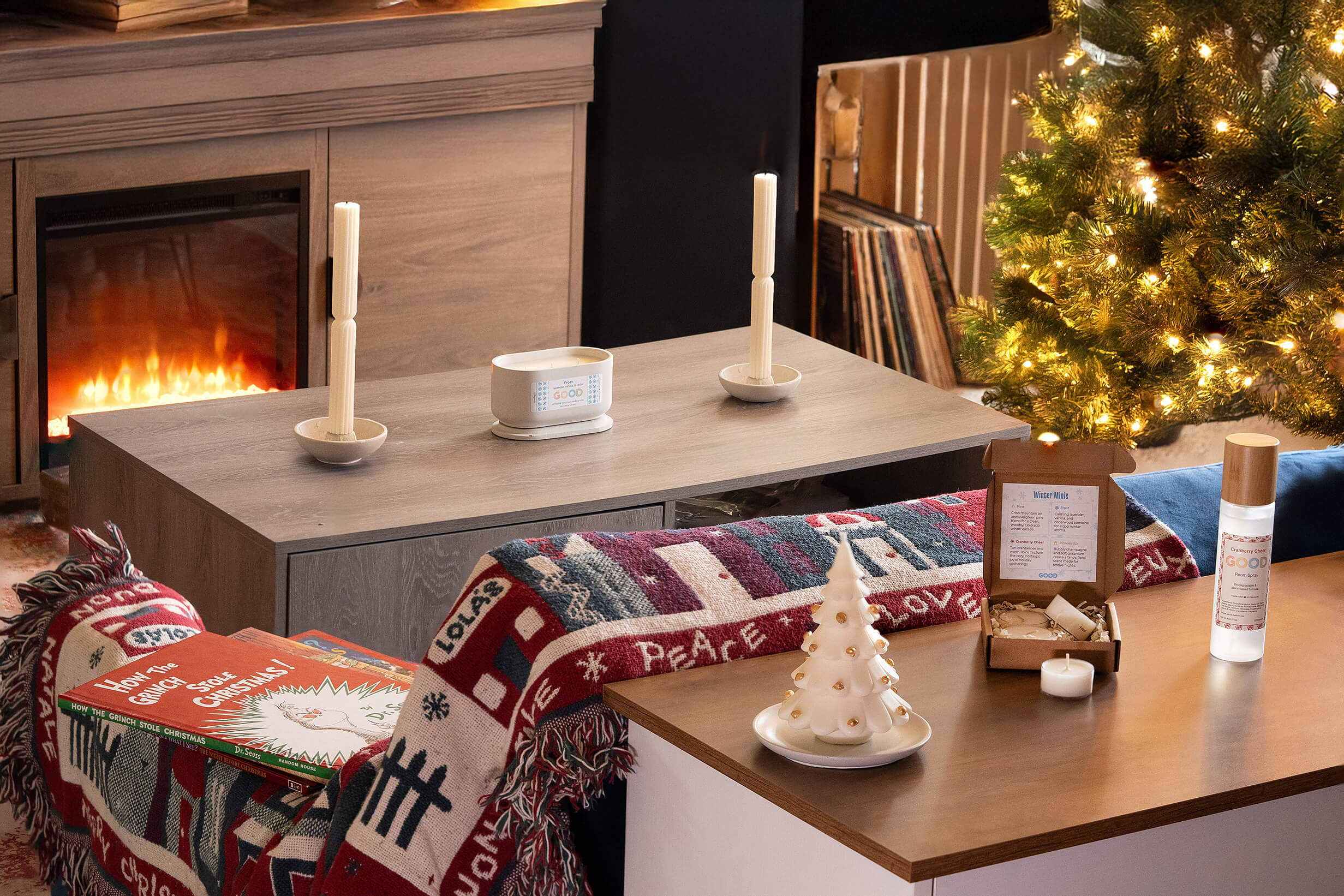 A cozy living room scene with a lit fireplace, Christmas tree, candles, books, and holiday decorations on tables, including a white ceramic tree, cookies, and a bottle of spray. A colorful blanket is draped over the couch.