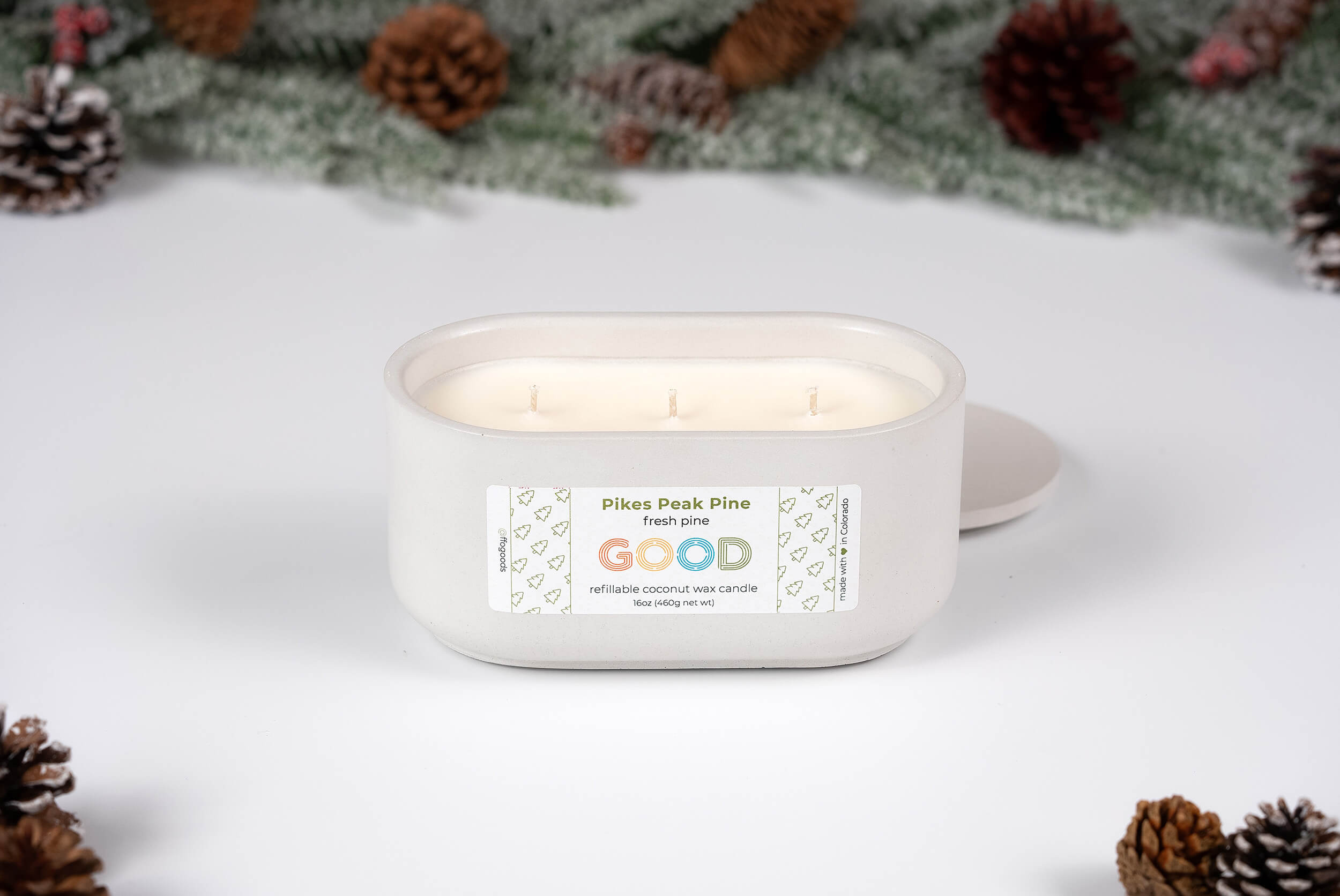 A white, oval “Pikes Peak Pine” three-wick candle is displayed on a white surface with its lid set aside, surrounded by pinecones and green foliage in the background.