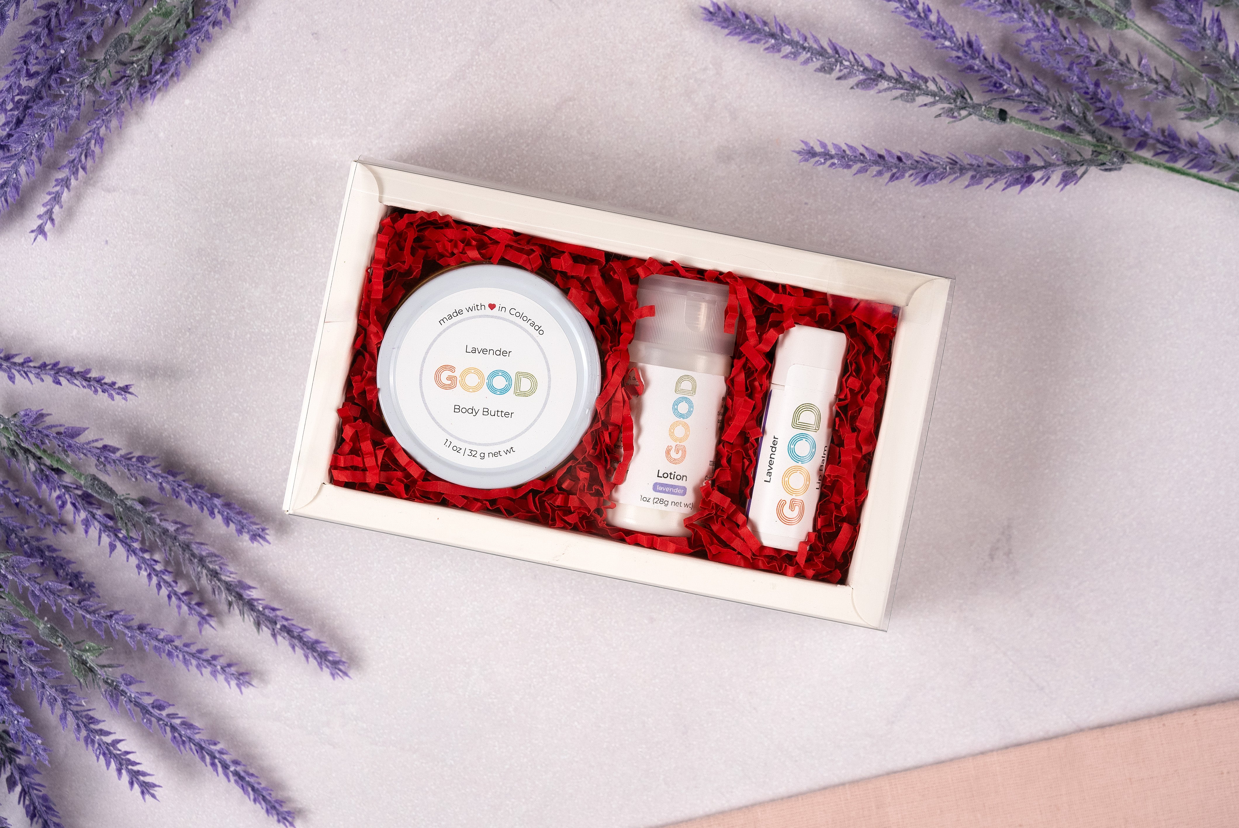 The Lavender Gift Set includes lavender body butter, lotion, and a GOOD-labeled Lavender Lip Balm, all arranged on a light surface with sprigs of lavender and red shredded paper filling the box.