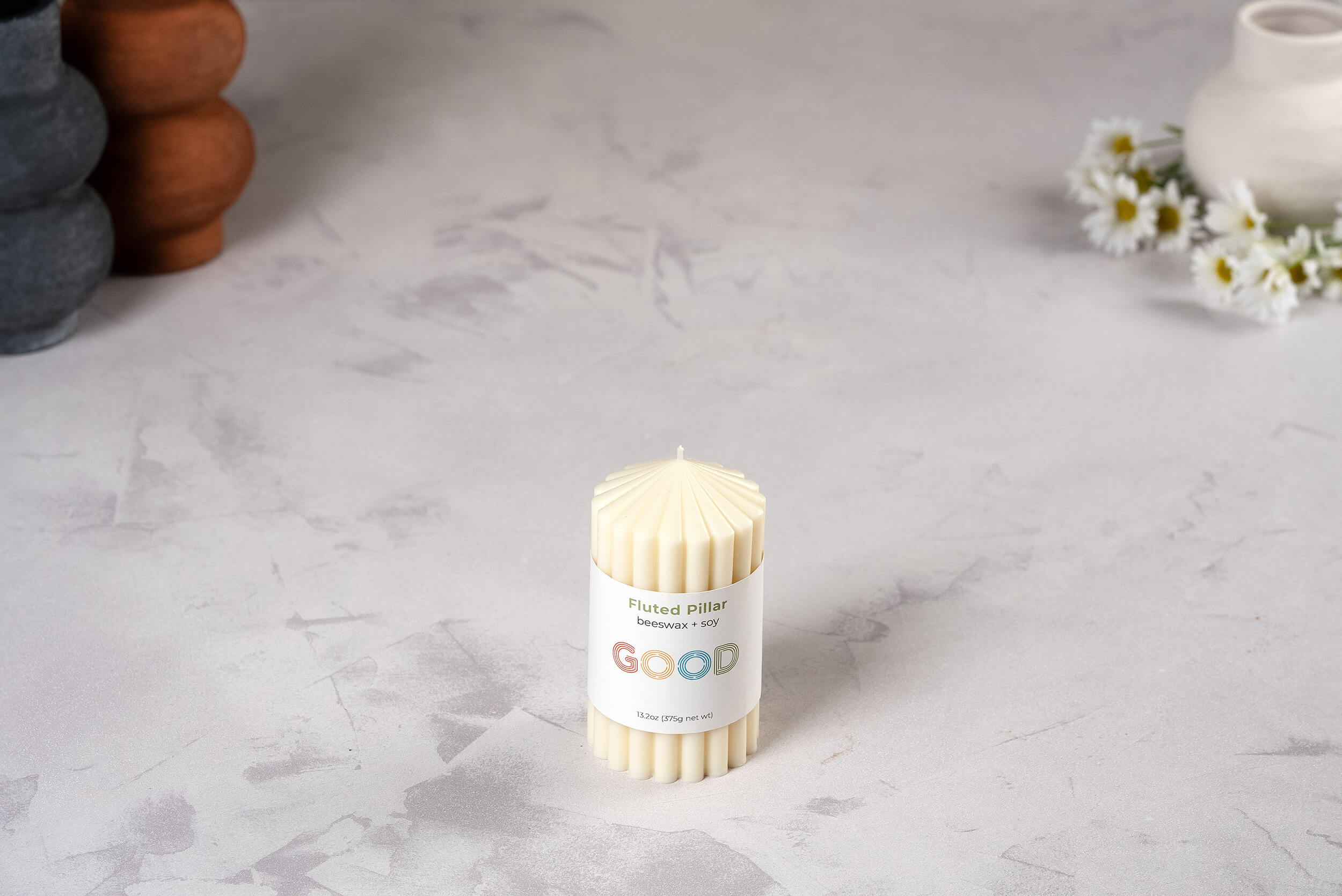 A cream fluted candle from our Fluted Pillar Candles set labeled "GOOD" rests on a light, textured surface, with white daisies and ceramic vessels in the background enhancing the charm of these elegant unscented candles.