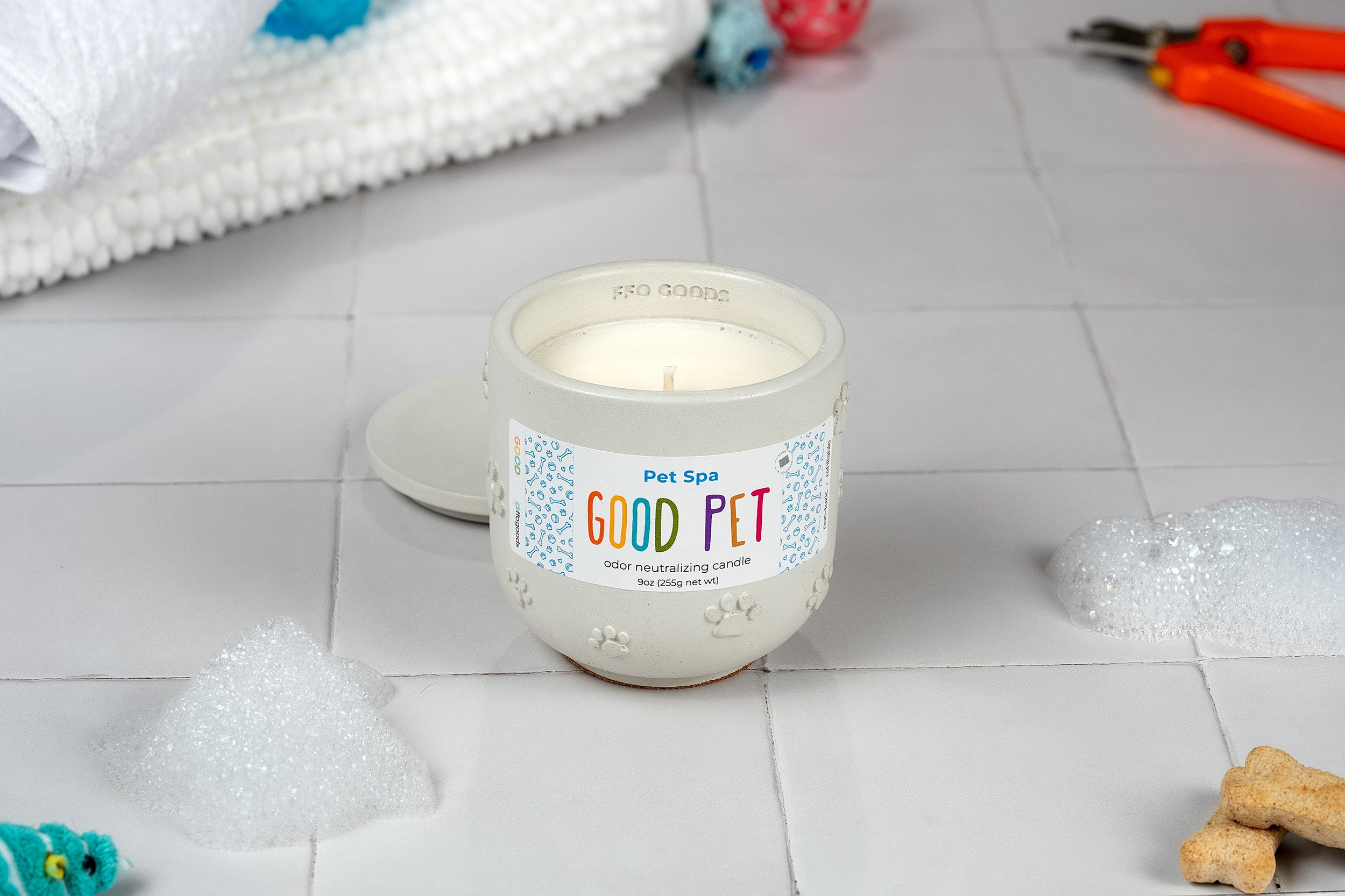 The Pet Spa candle, in a refillable concrete vessel, rests on white tiles with bubbles, a towel, pet treats, and grooming tools nearby, providing an odor-eliminating scent for a relaxing pet spa vibe.