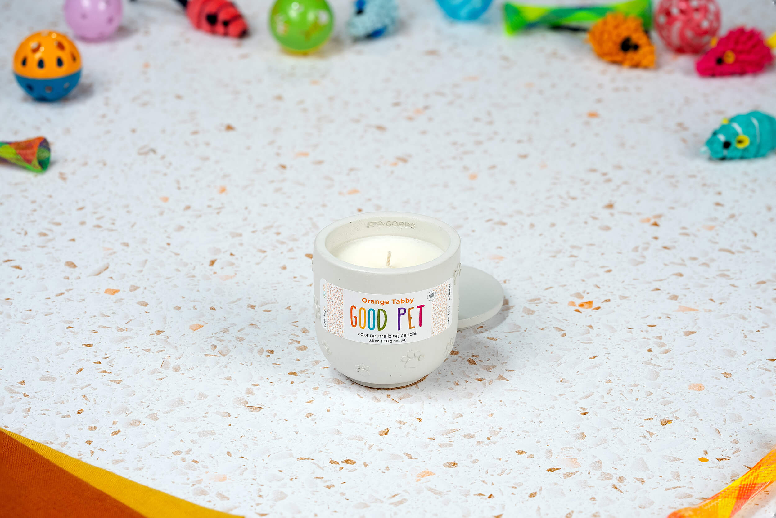 An Orange Tabby Good Pet odor-eliminating candle in a small mug rests on a terrazzo surface, with colorful cat toys in the background.