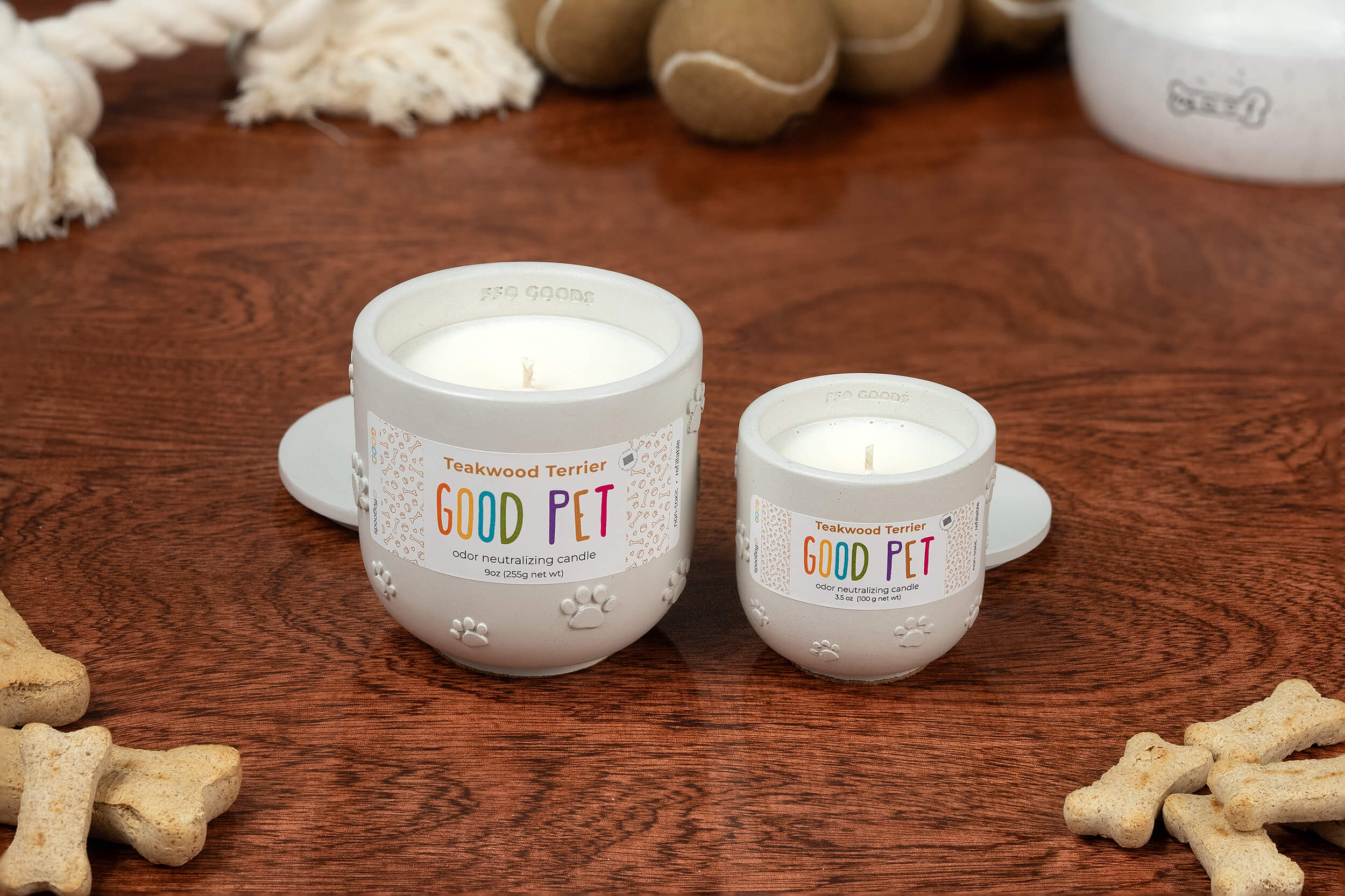 Two white Teakwood Terrier candles labeled GOOD PET, with paw prints and odor-neutralizing scents, rest on a wooden surface among dog biscuits, toys, and a white bowl—a great choice for pet lovers wanting fresher air.