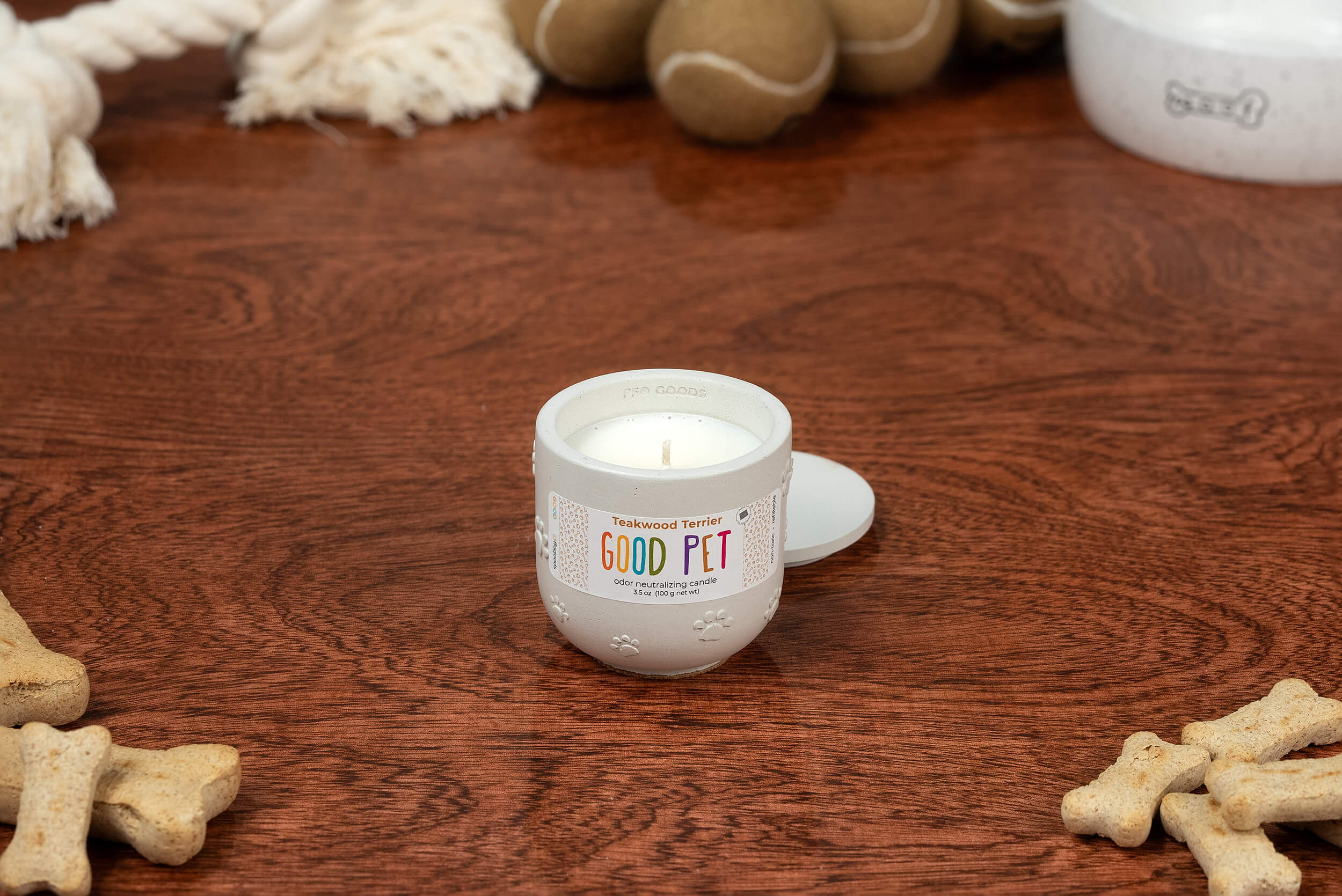 A Teakwood Terrier candle in a small jar labeled “GOOD PET” sits open on wood, surrounded by dog toys, rope, and bone treats—ready to release its odor-eliminating scent.