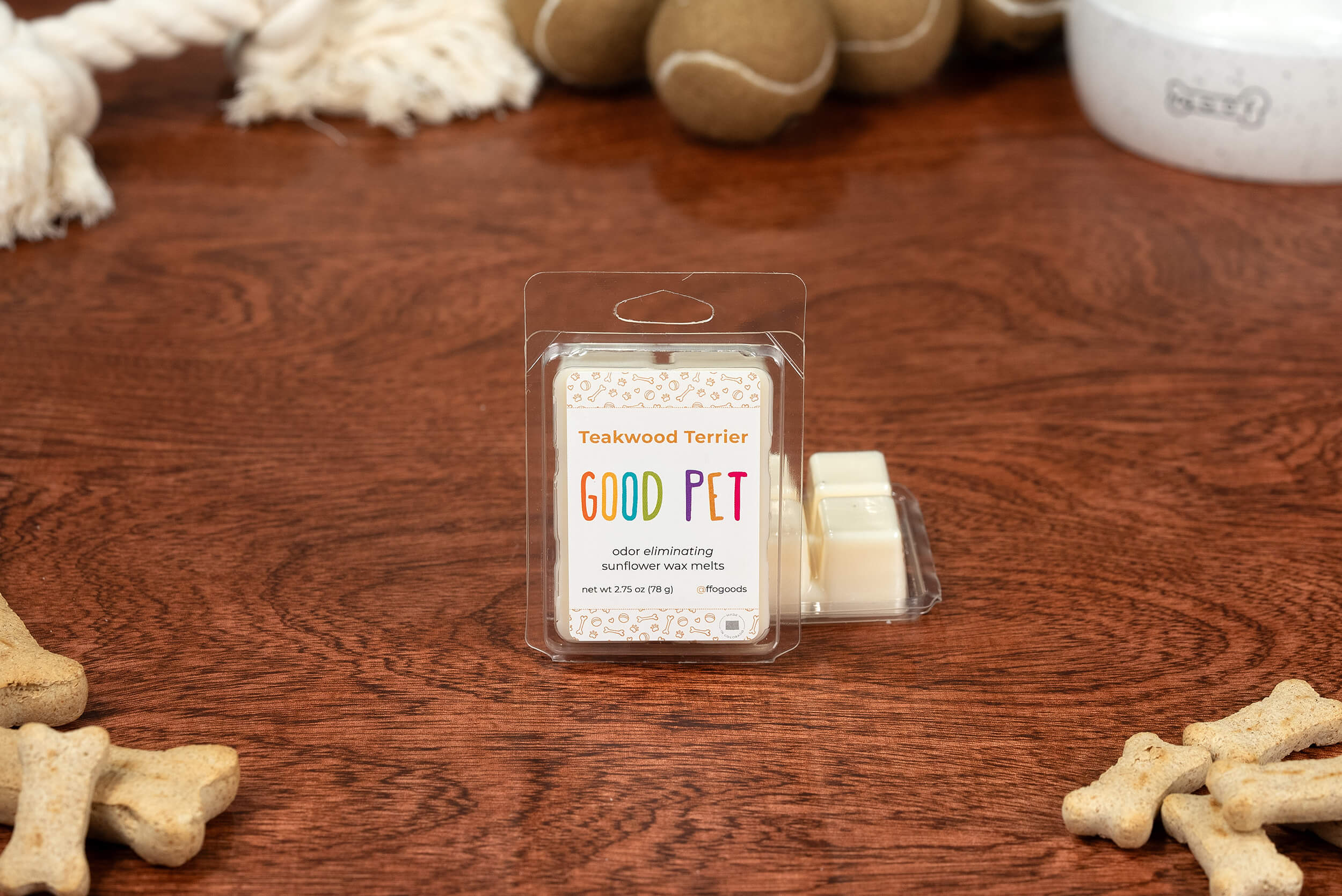 A package of Teakwood Terrier Wax Melts is on a wooden table, surrounded by dog toys, bones, and a pet bowl. These plant-based wax melts freshen your space with odor-eliminating scents.