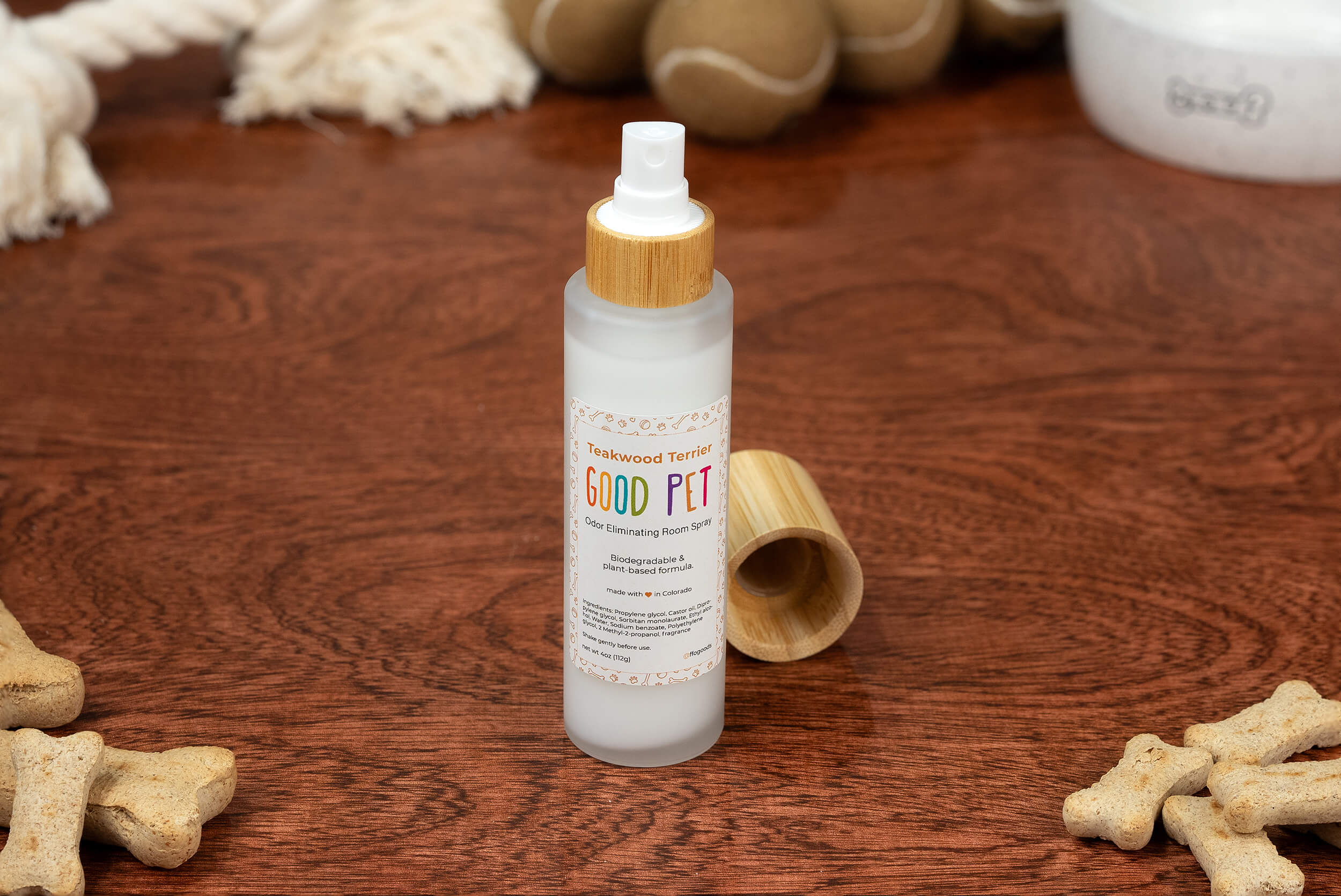 A bottle of Good Pet Room Spray with a wooden cap rests on a wooden surface, surrounded by dog bones, rope toys, and a white pet bowl—ideal to pair with your favorite plant-based air freshener or biodegradable odor eliminator.
