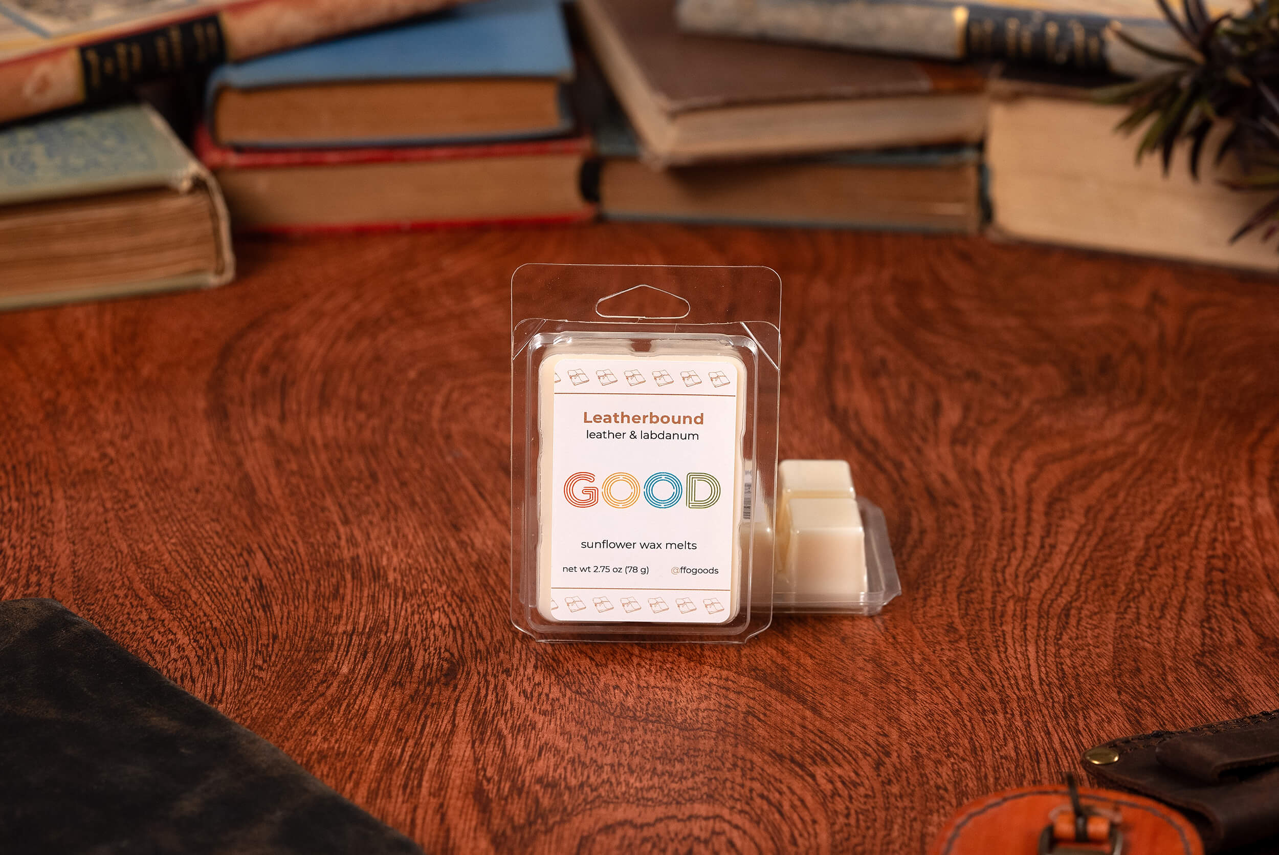 A package of Leatherbound sunflower wax melts rests on a wooden table among old books, a plant, and a black notebook. Two wax cubes are separated from the pack, accentuating the warm leather scent.