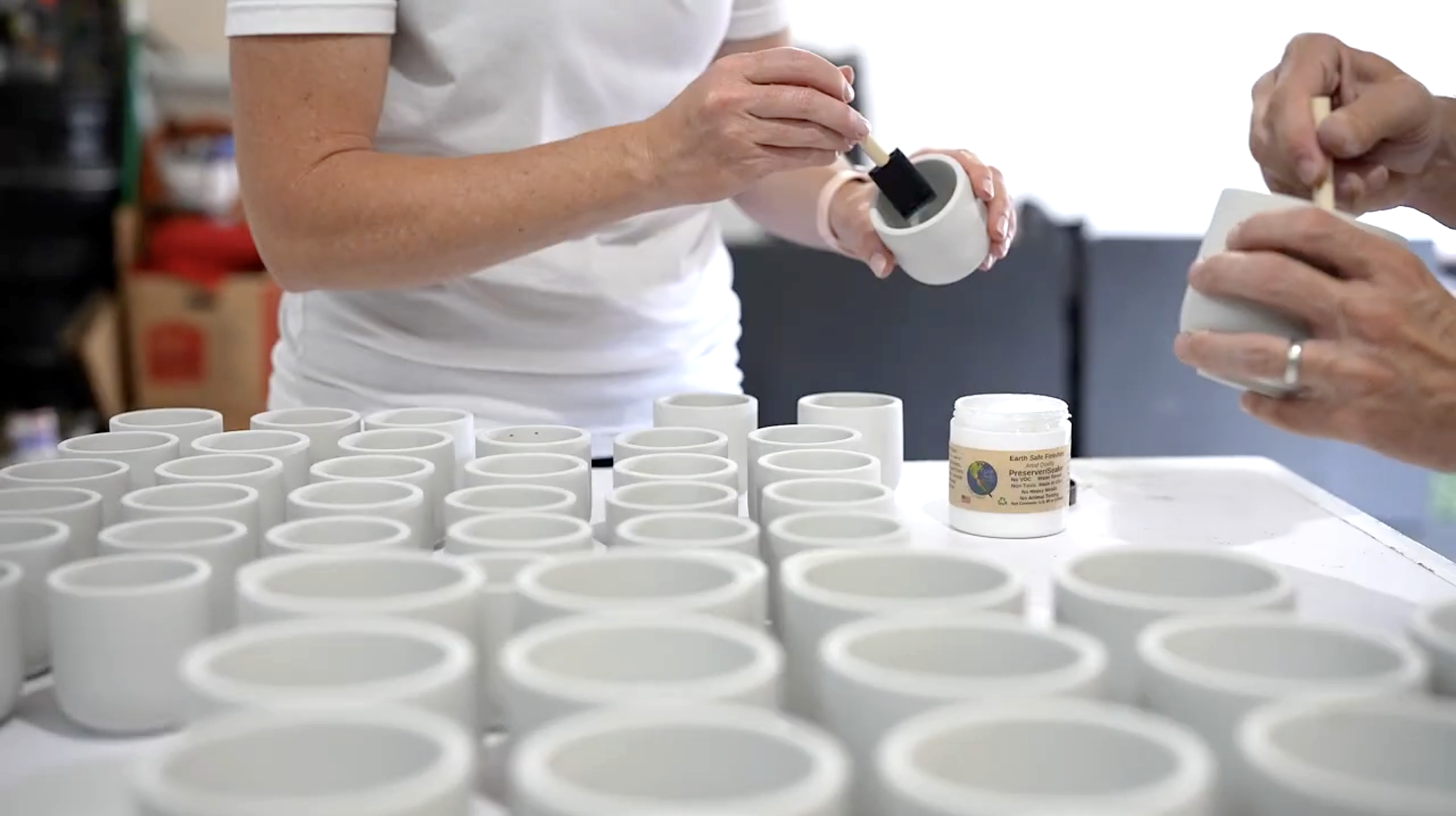 A person in a white shirt paints the inside of a small white ceramic cup with black paint at a table full of similar unpainted cups. Another hand holds a cup on the right.