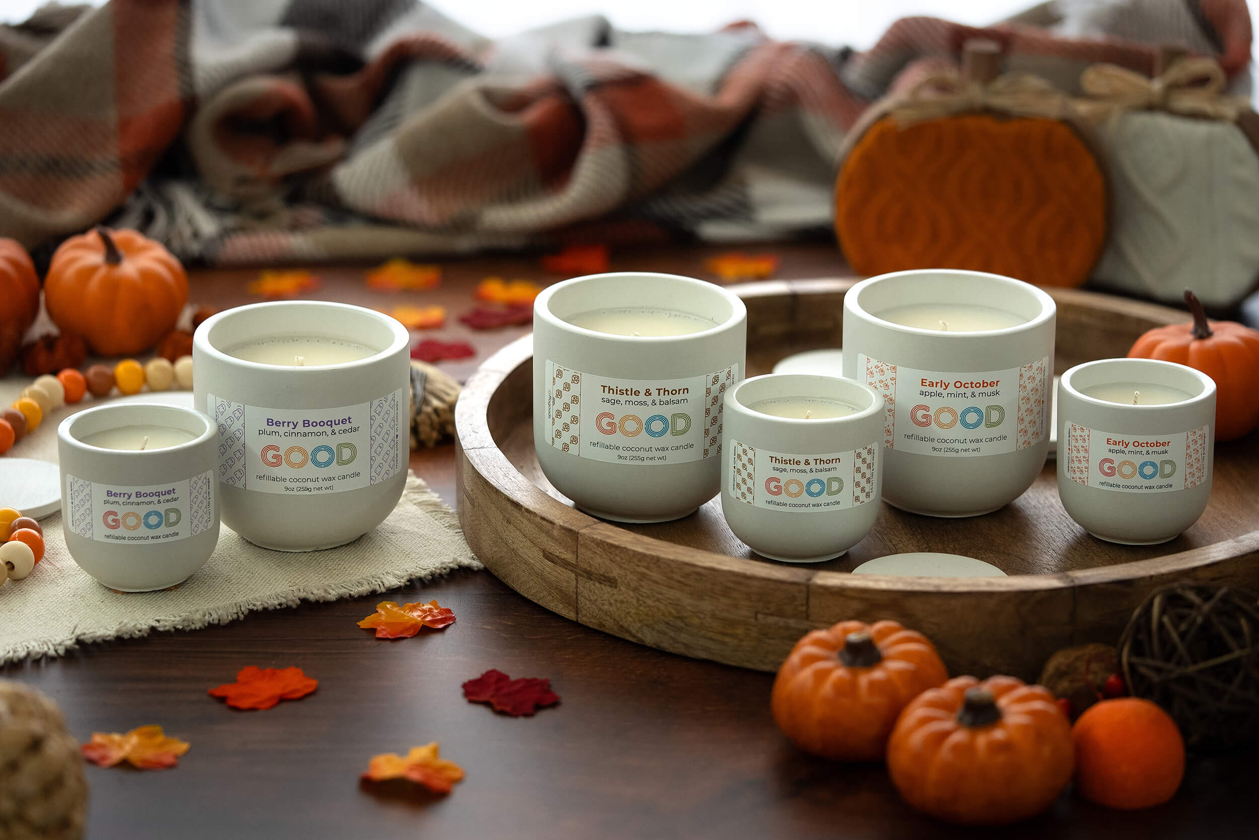 Five white candles with colorful labels sit on a wooden tray surrounded by small pumpkins, fall leaves, beads, and a cozy plaid blanket, creating a warm autumn atmosphere.