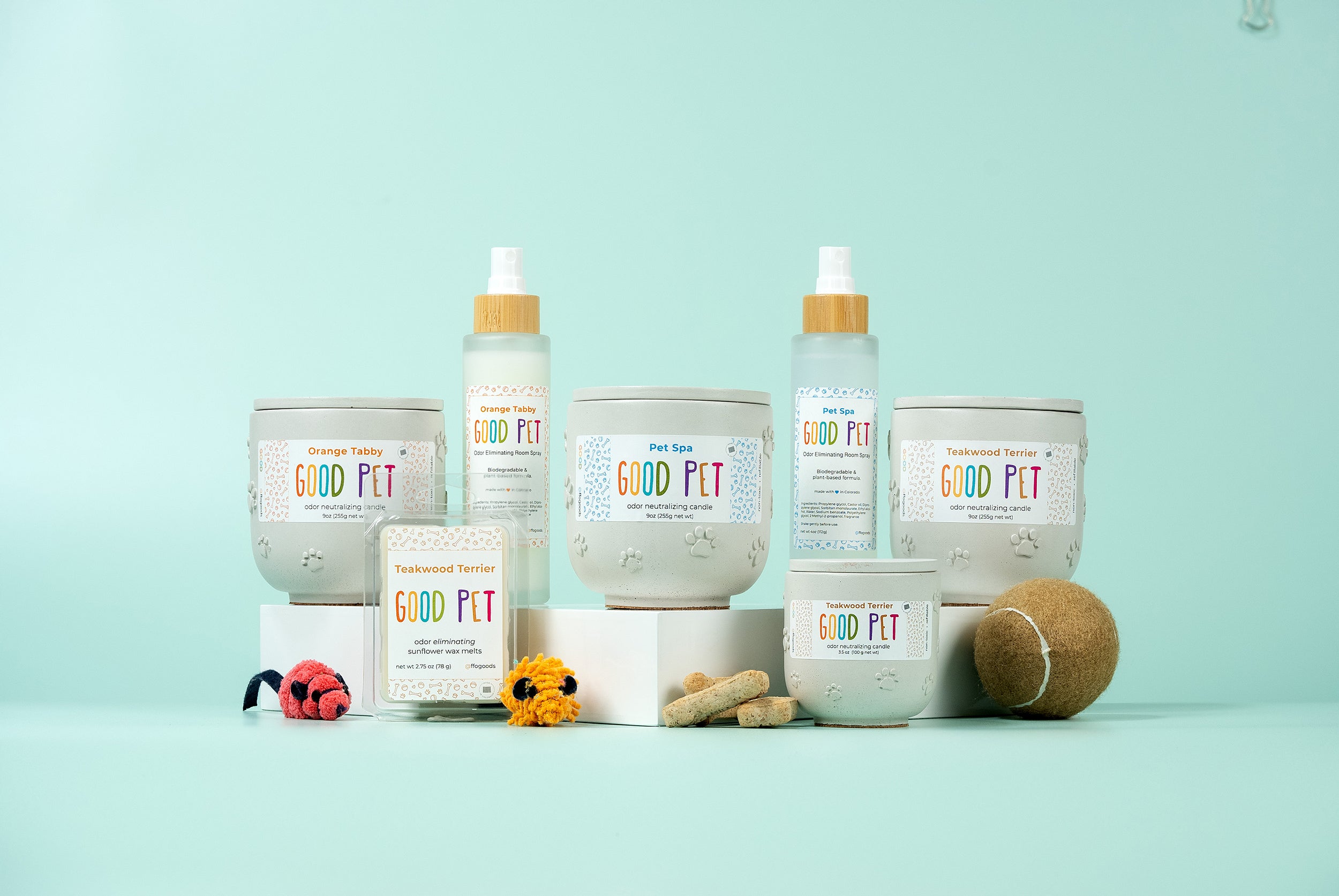 Several GOOD PET grooming products and treats are arranged on white stands against a light blue background, along with pet toys and a tennis ball in the foreground.