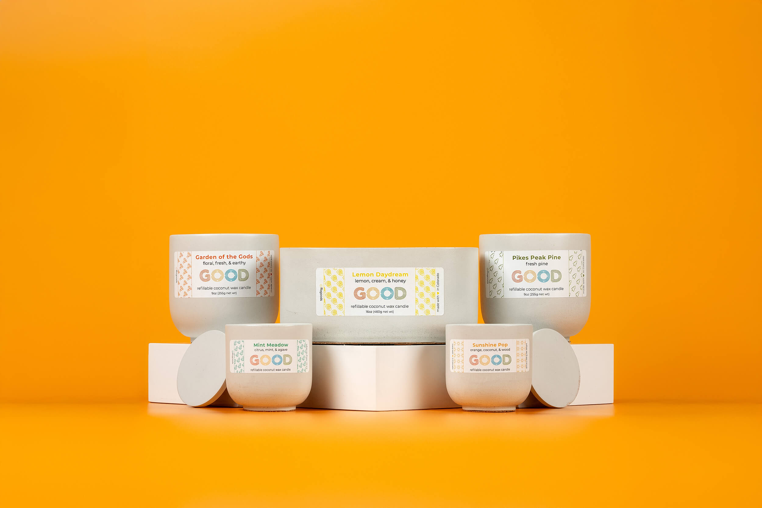 Five white candle jars with colorful labels sit on white stands against a vibrant orange background. Each label displays the brand GOOD and different scent names.