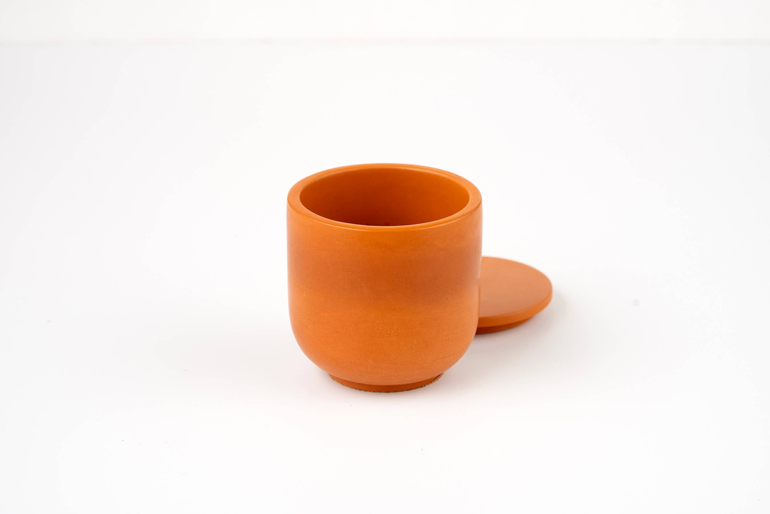 A small, round, orange clay Orange Lilly Candle Vessel with a matching flat lid resting behind it, displayed on a plain white surface.