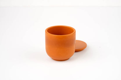 A small, round, orange clay Orange Lilly Candle Vessel with a matching flat lid resting behind it, displayed on a plain white surface.