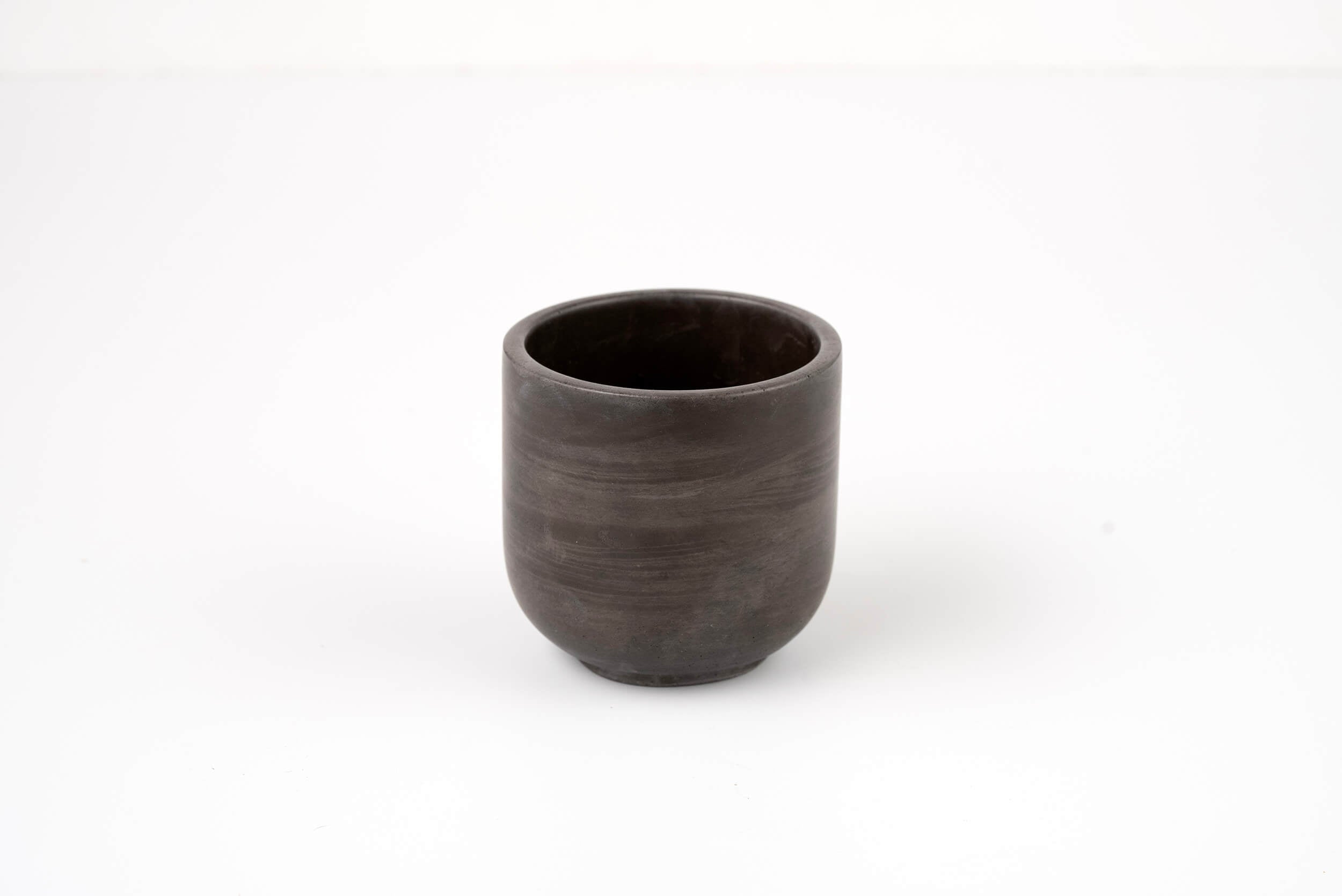 The Graphite Candle Vessel, with its simple dark ceramic design and smooth, rounded shape, rests on a plain white surface and evokes a minimalist aesthetic when viewed from the front.