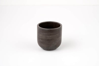 The Graphite Candle Vessel, with its simple dark ceramic design and smooth, rounded shape, rests on a plain white surface and evokes a minimalist aesthetic when viewed from the front.