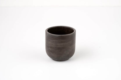 The Graphite Candle Vessel, with its simple dark ceramic design and smooth, rounded shape, rests on a plain white surface and evokes a minimalist aesthetic when viewed from the front.