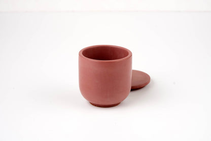The Antique Rose Candle Vessel features a simple, round, reddish-brown design with a matching lid displayed beside it on a white background.