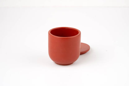 A small, round, red Lava Candle Vessel with a matching lid sits beside it on a plain white background.
