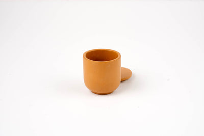 The Wheat Buff Candle Vessel, a small round brown clay cup with a matching saucer behind it, is displayed against a plain white background.